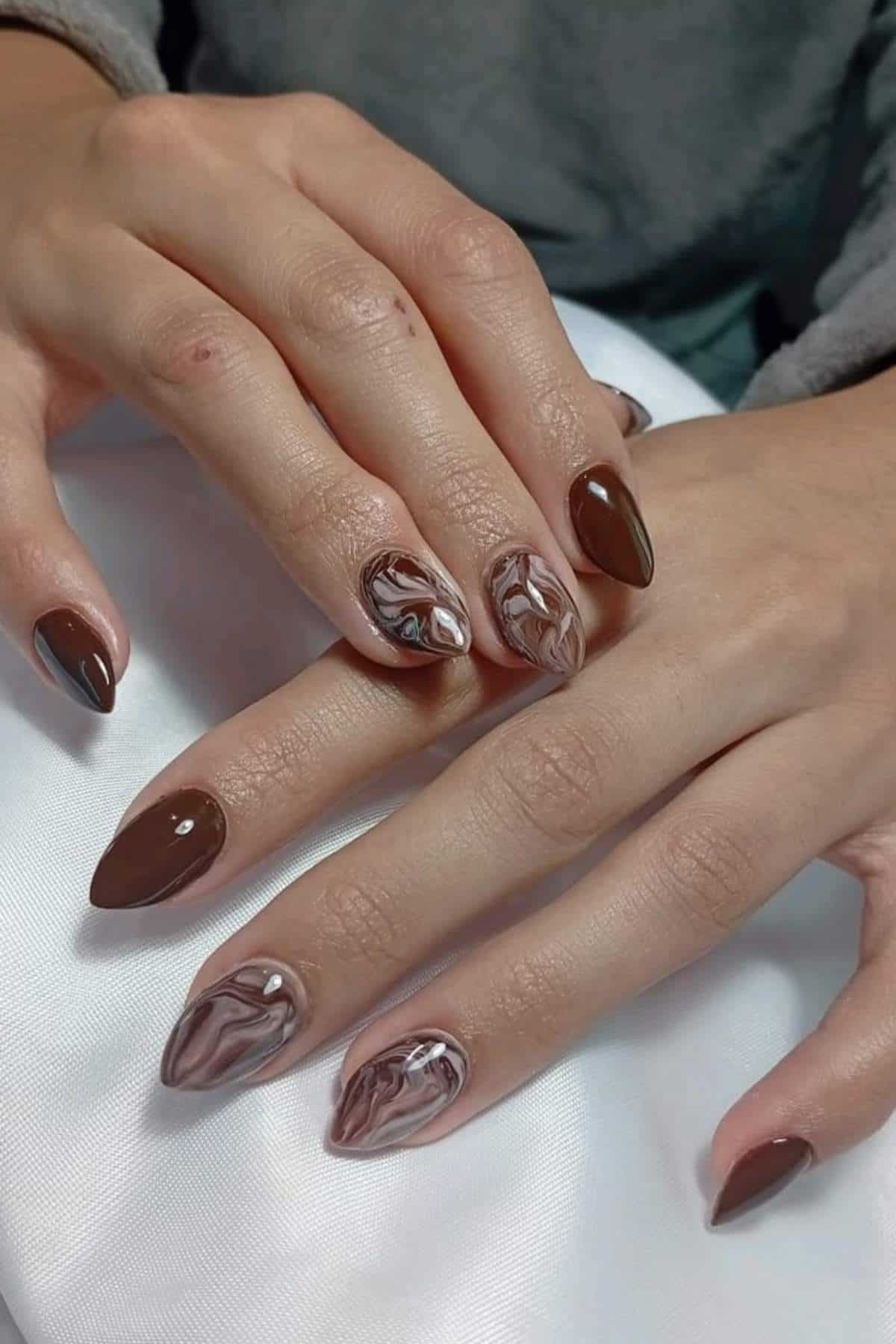 Hands with almond-shaped nails featuring a brown and beige marbled design on some nails and solid brown polish on others, resting on a white surface.