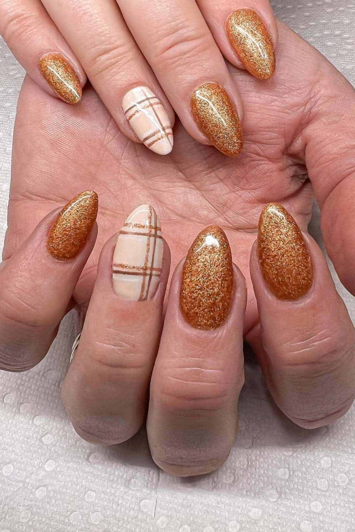 A hand with almond-shaped nails; most are painted with shimmery bronze polish, while two have a beige base with brown and gold plaid designs.