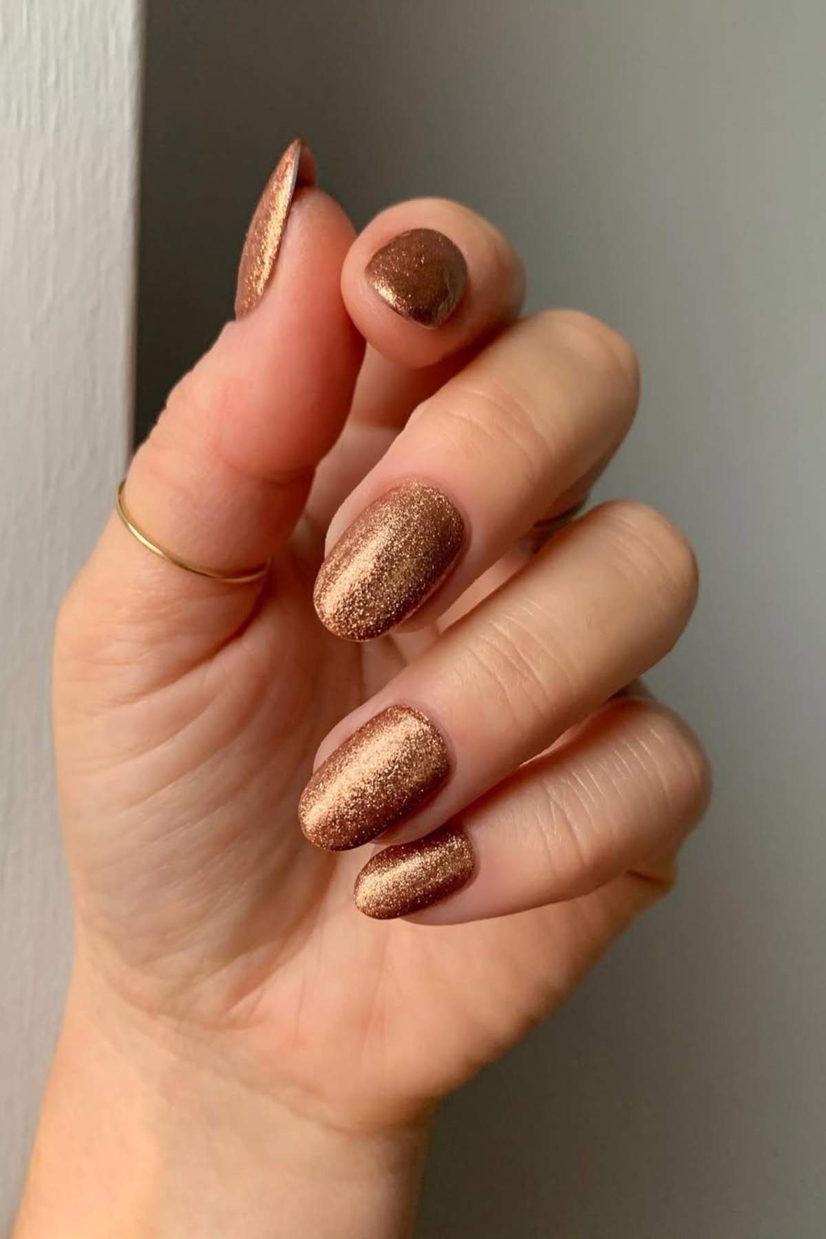 A hand with short, oval-shaped fingernails painted in shimmery bronze nail polish, and a thin gold ring on the middle finger.