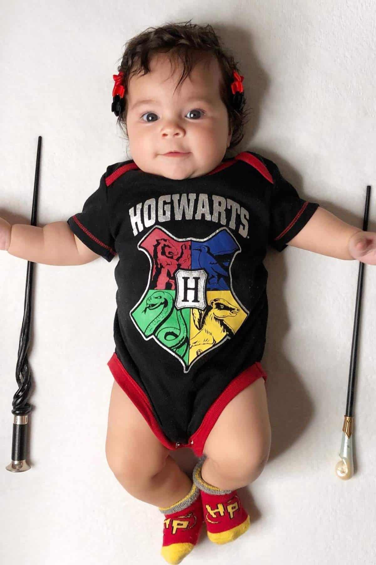 Baby lying on a white surface, wearing a Hogwarts-themed onesie and red-and-yellow socks, with two wands placed on either side.