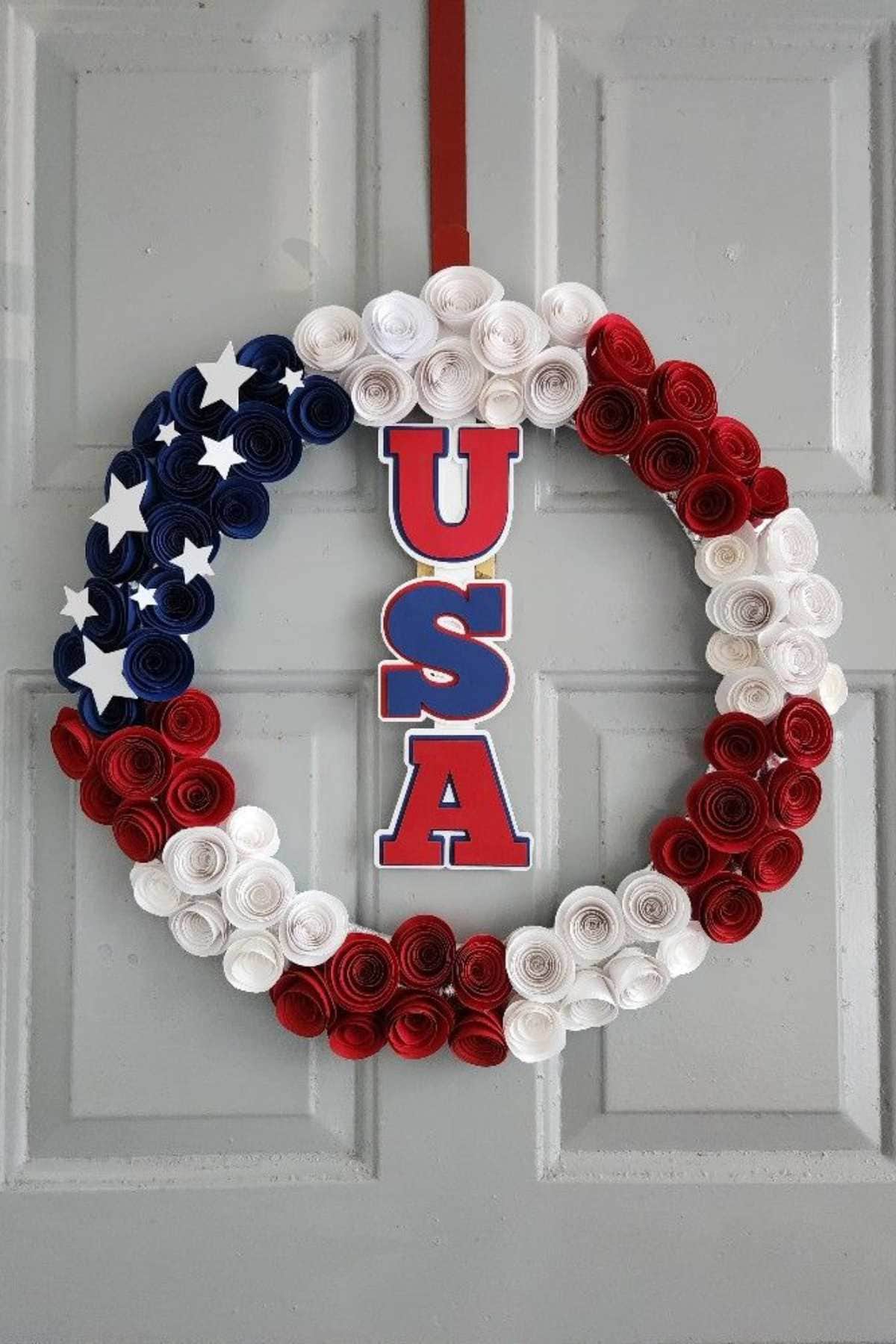 A wreath made of red, white, and blue paper flowers with white stars and a vertical "USA" sign hangs on a gray door.