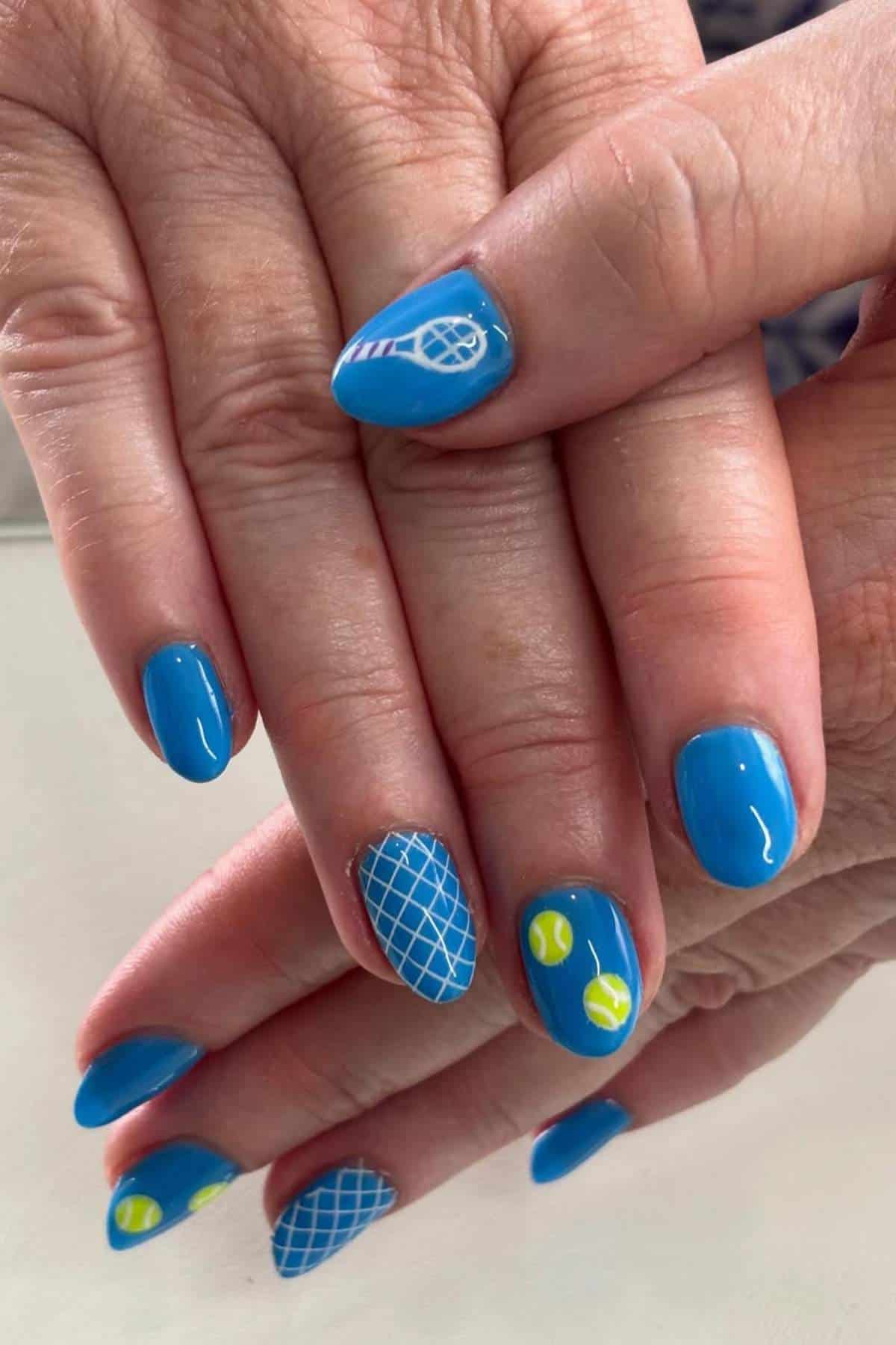 Hands with blue painted fingernails featuring white tennis racket and net designs, and small yellow tennis balls as nail art accents.