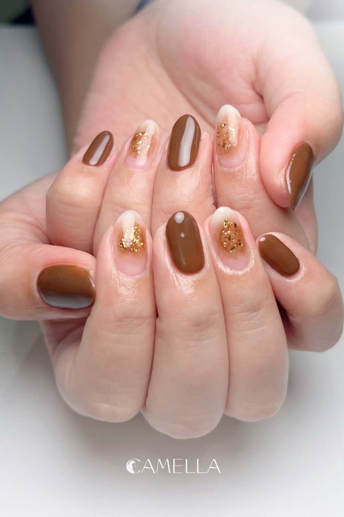 Hands with manicured nails featuring brown polish and accent nails with clear base and gold glitter.