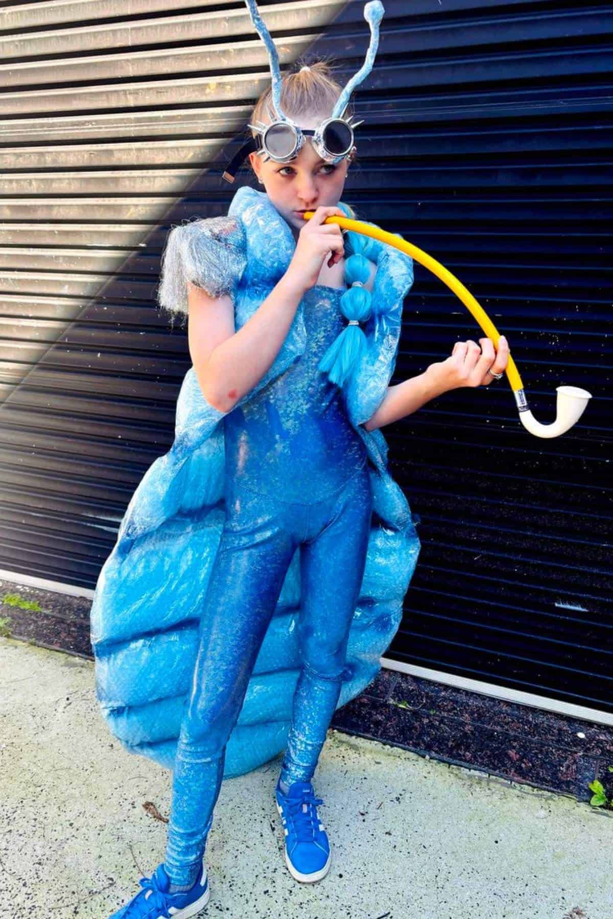 Child in a shiny blue caterpillar costume with antennae, wearing goggles, and holding a yellow pipe, standing in front of a black corrugated wall.