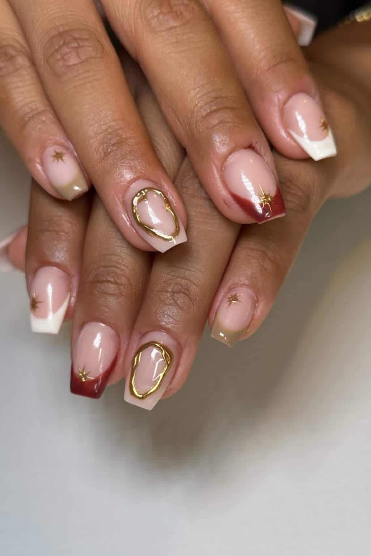 Hands with square-shaped nails featuring a nude base, white and burgundy French tips, gold star accents, and gold abstract line designs on two nails.