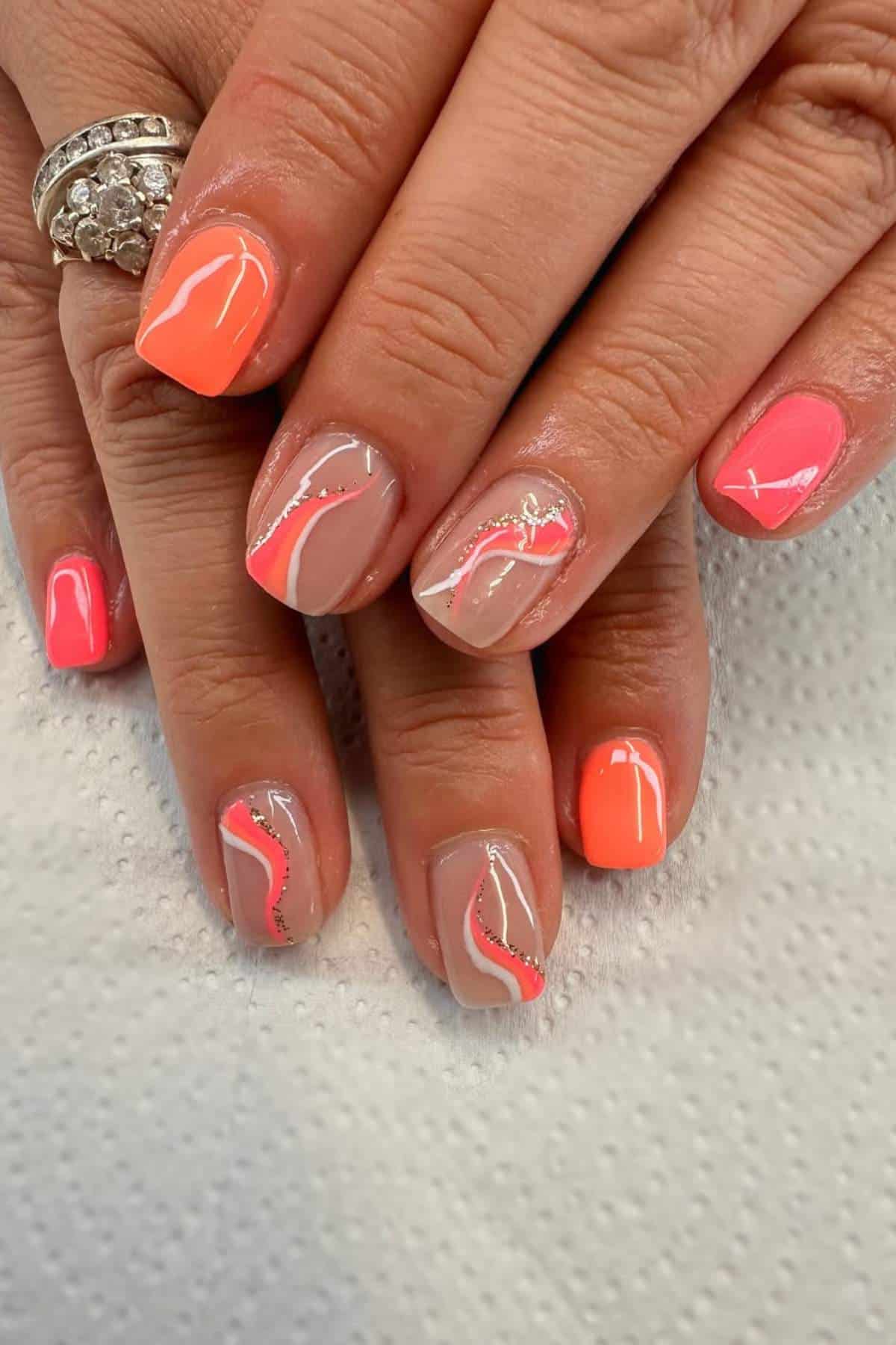 Hands with short, square-shaped nails featuring a mix of bright coral polish and nude base with pink, white, and silver wavy line designs. Rings are worn on two fingers.