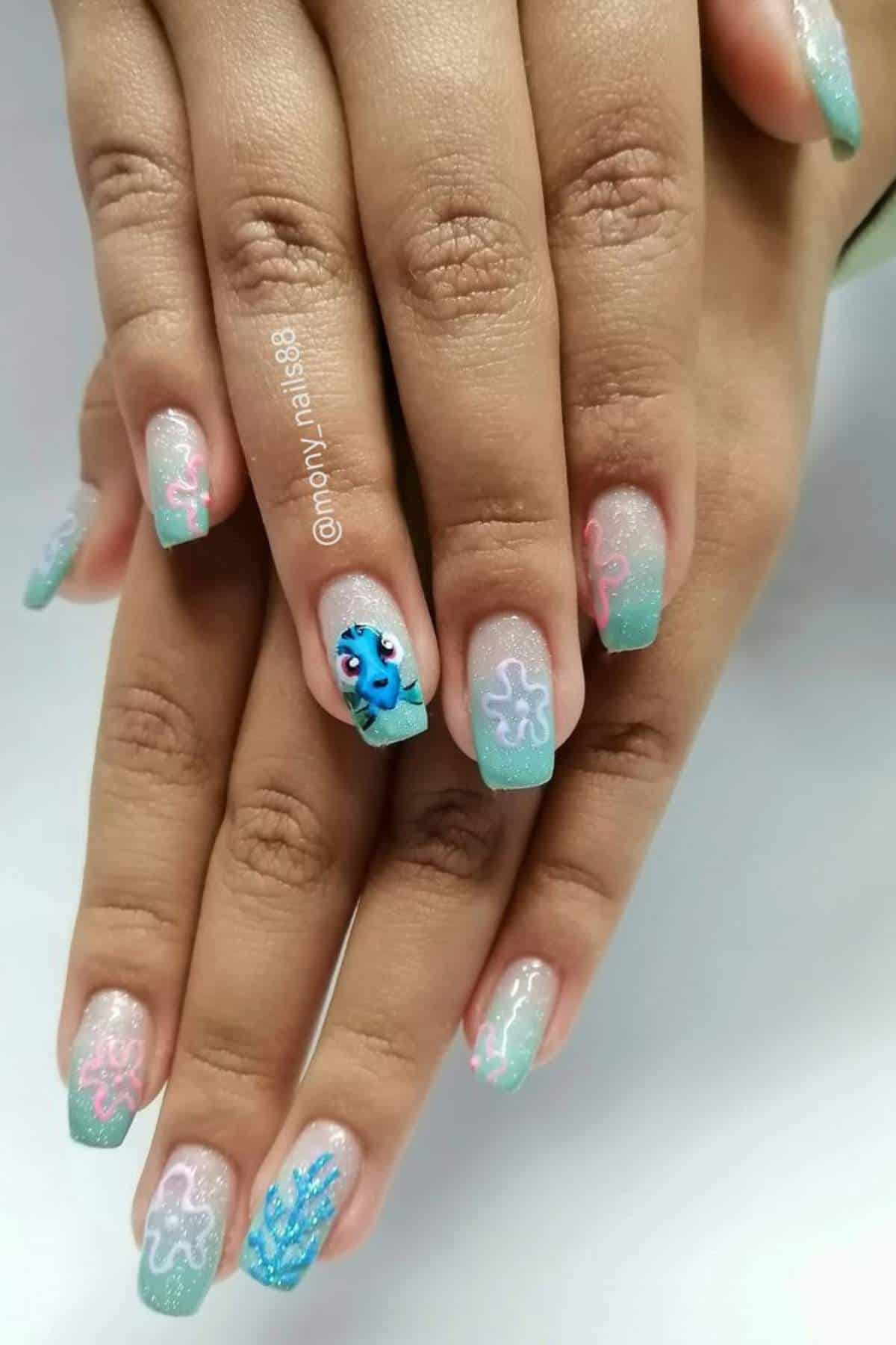 Hands with long square acrylic nails featuring turquoise and pink abstract designs, glitter accents, and a blue cartoon character on the ring finger.