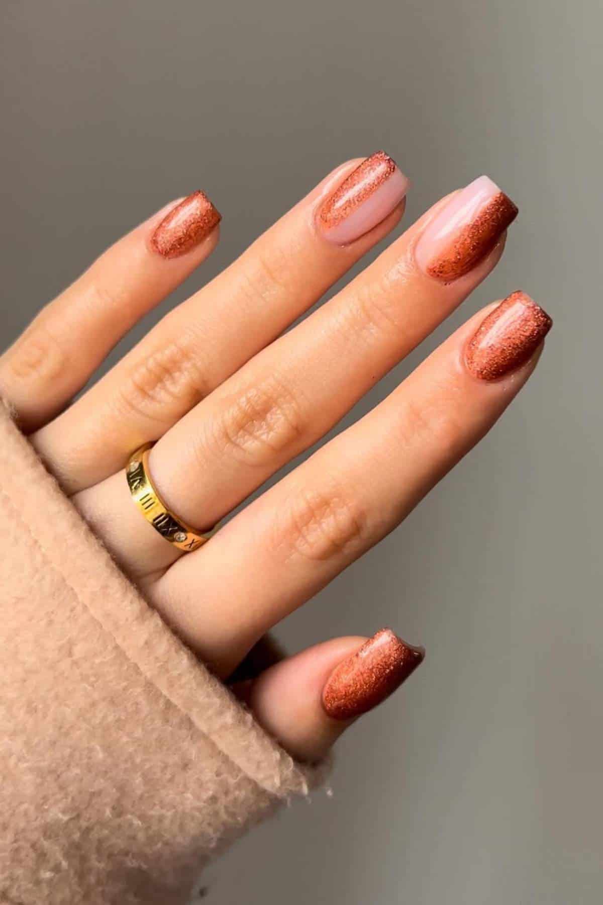 A hand with short, square-shaped nails painted with a brown glittery polish and a gold ring on the ring finger. The sleeve of a beige coat covers part of the hand.