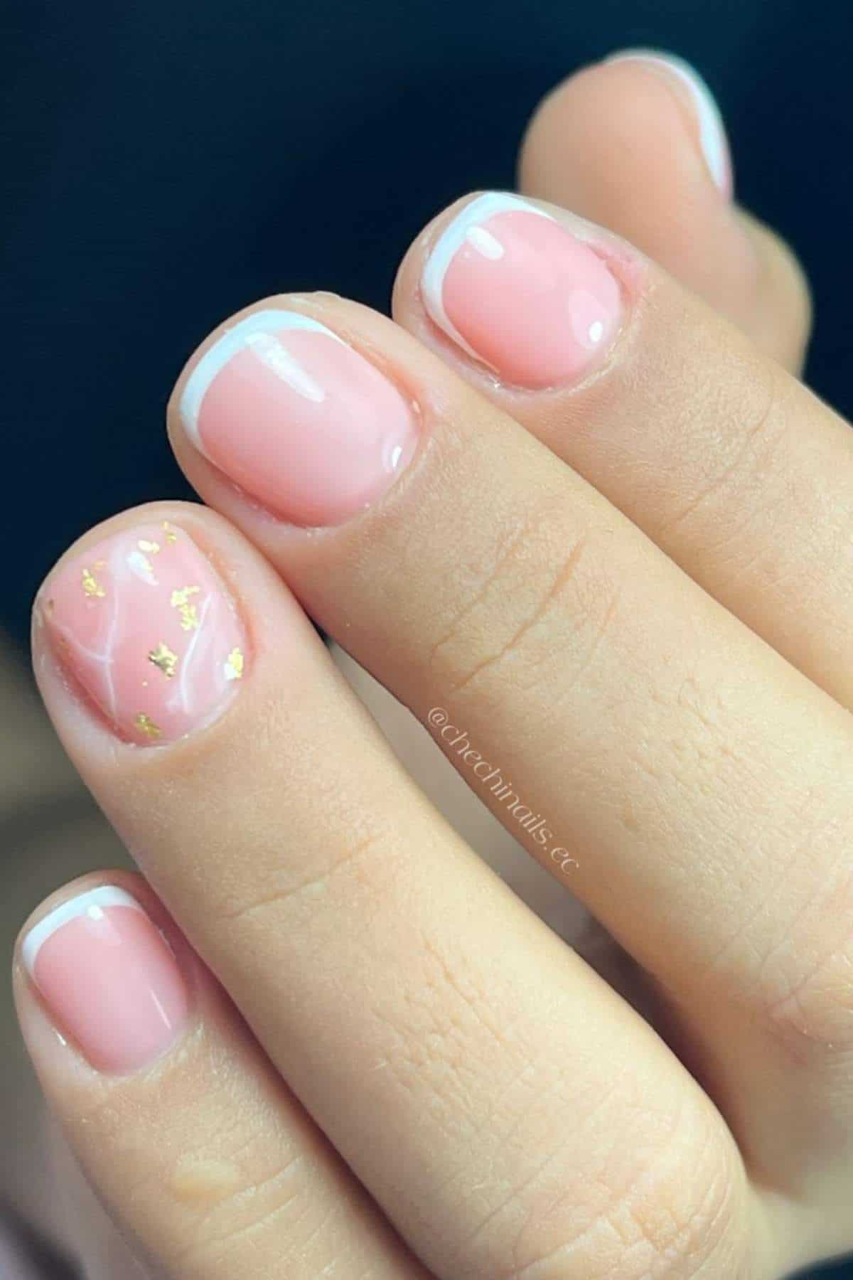 A hand with short, rounded nails painted with a light pink polish, white French tips, and gold foil accents on the ring finger.