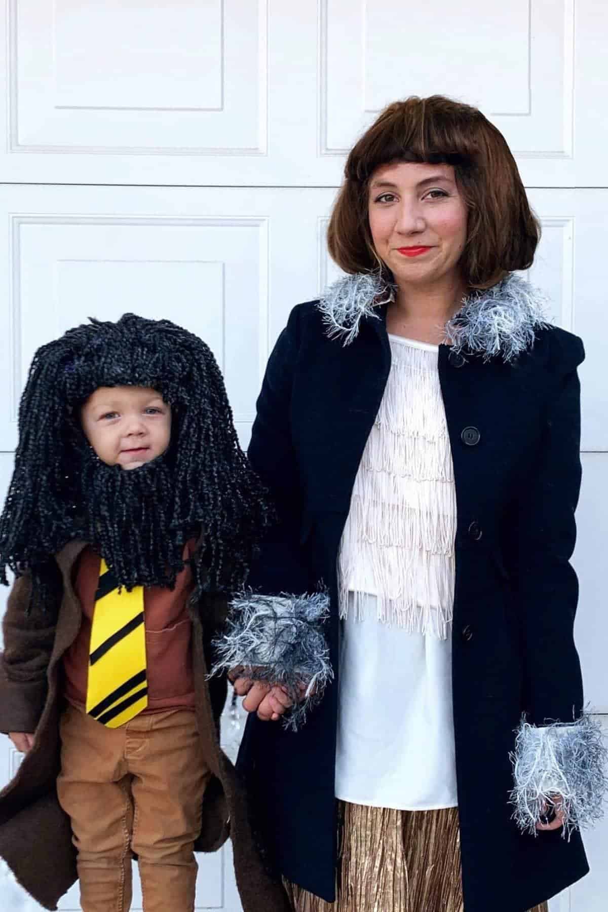 An adult and a child stand together in costumes, with the adult wearing a dark coat and wig, and the child wearing a wig, brown coat, and yellow-striped tie.