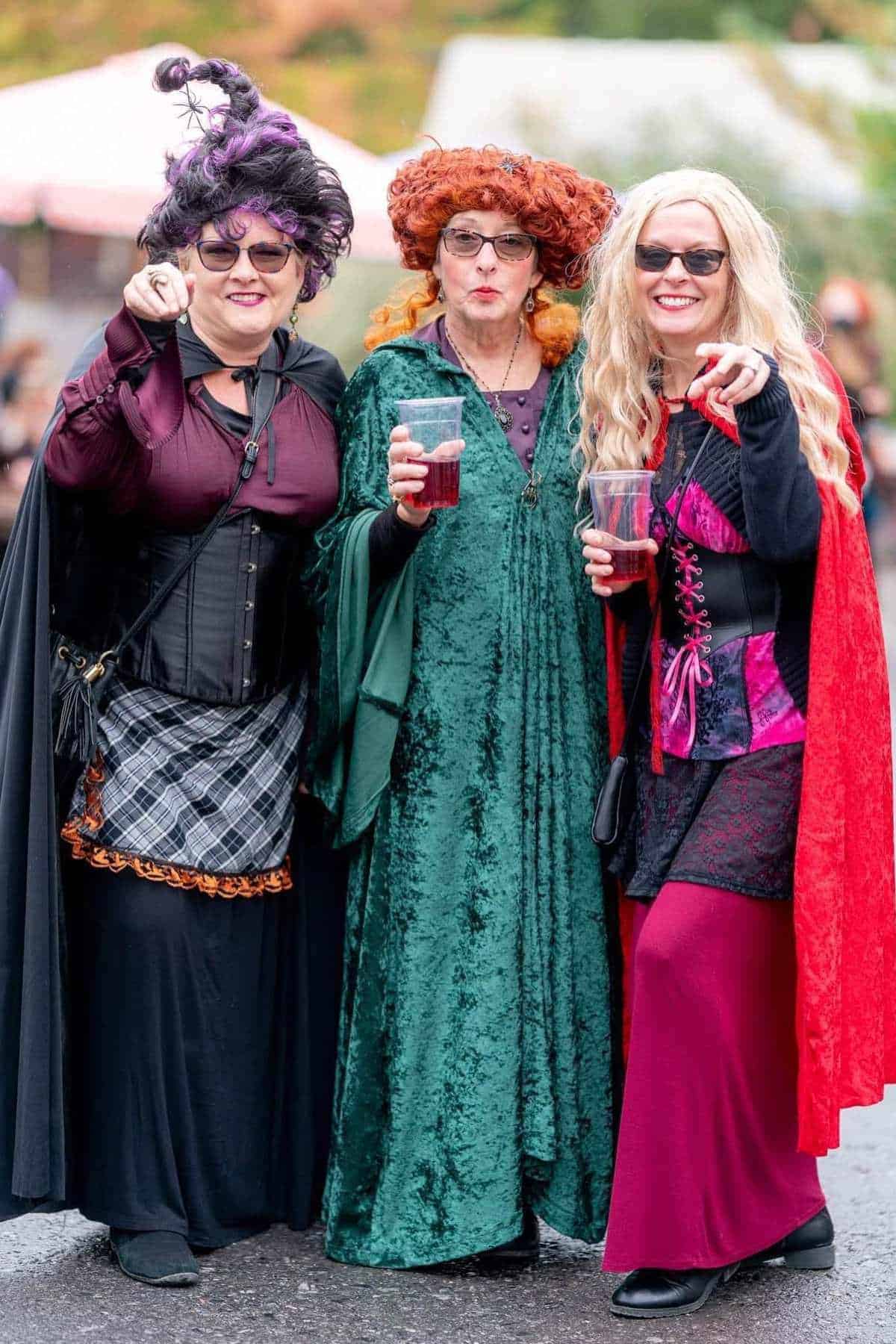 Three women in colorful witch costumes and sunglasses stand together outdoors, holding drinks and smiling at the camera.
