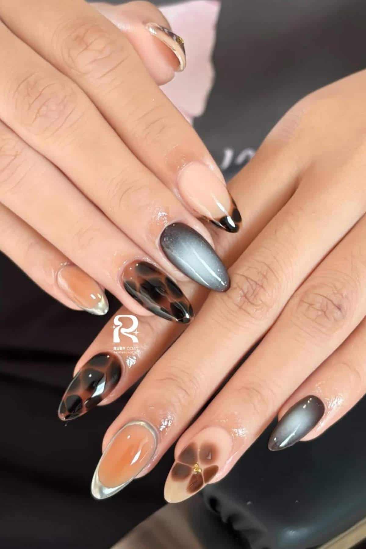 Hands with almond-shaped nails featuring a mix of black, nude, metallic gray, and brown abstract designs and patterns on each nail.