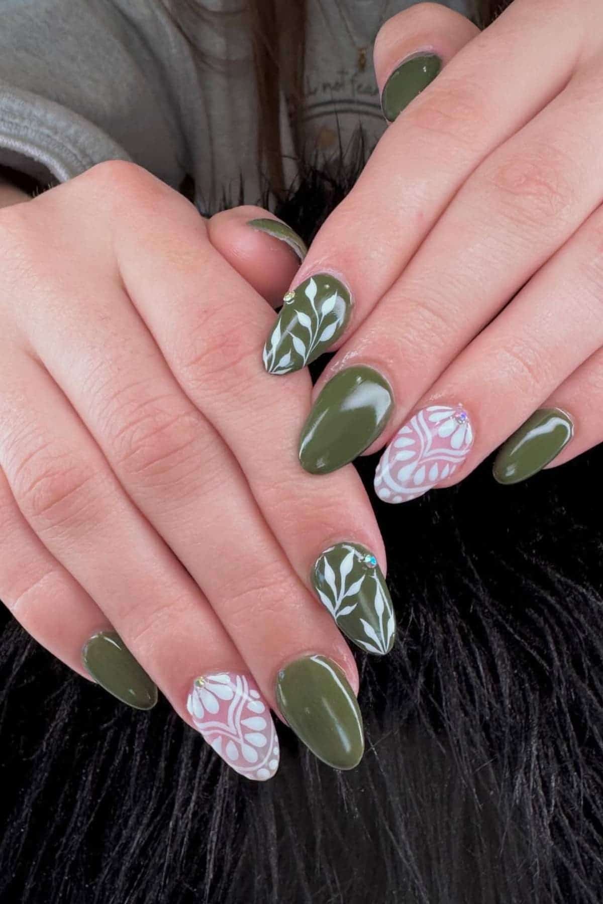 Hands with long, almond-shaped nails painted dark green, with accent nails featuring white and pink floral and leafy designs. The hands rest on a black textured surface.