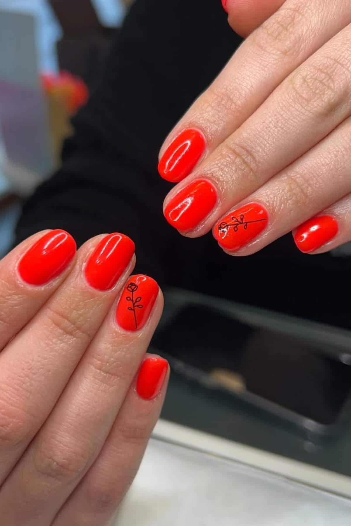 Close-up of hands with glossy red nail polish; two nails feature a small black botanical line design.