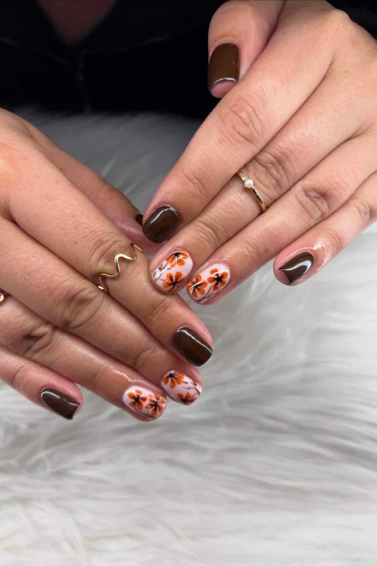 Hands with brown and nude nails, some featuring orange and black floral designs. The fingers have gold rings and are resting on a white textured surface.