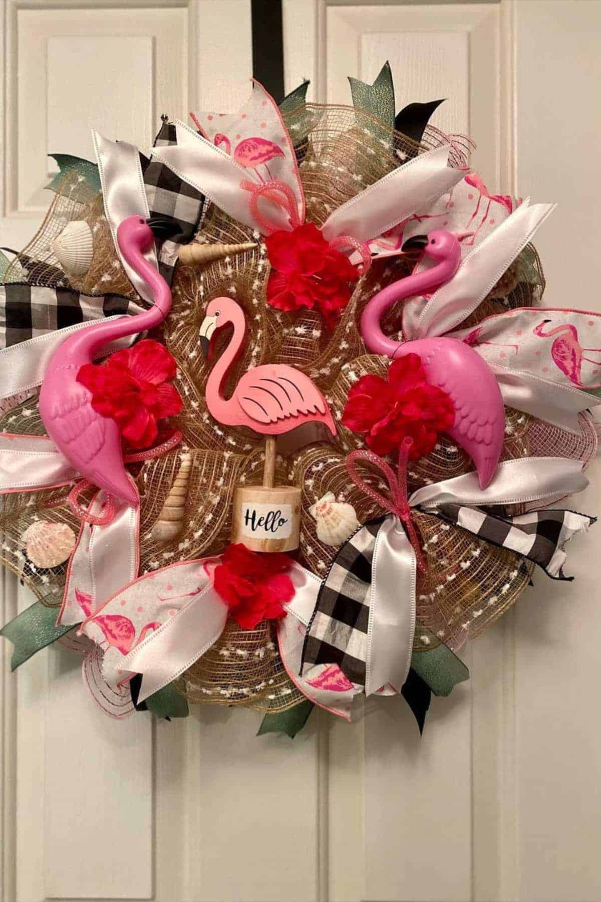 A decorative wreath featuring three pink flamingos, red flowers, seashells, and black-and-white ribbon bows, with a small sign that says "Hello" in the center.