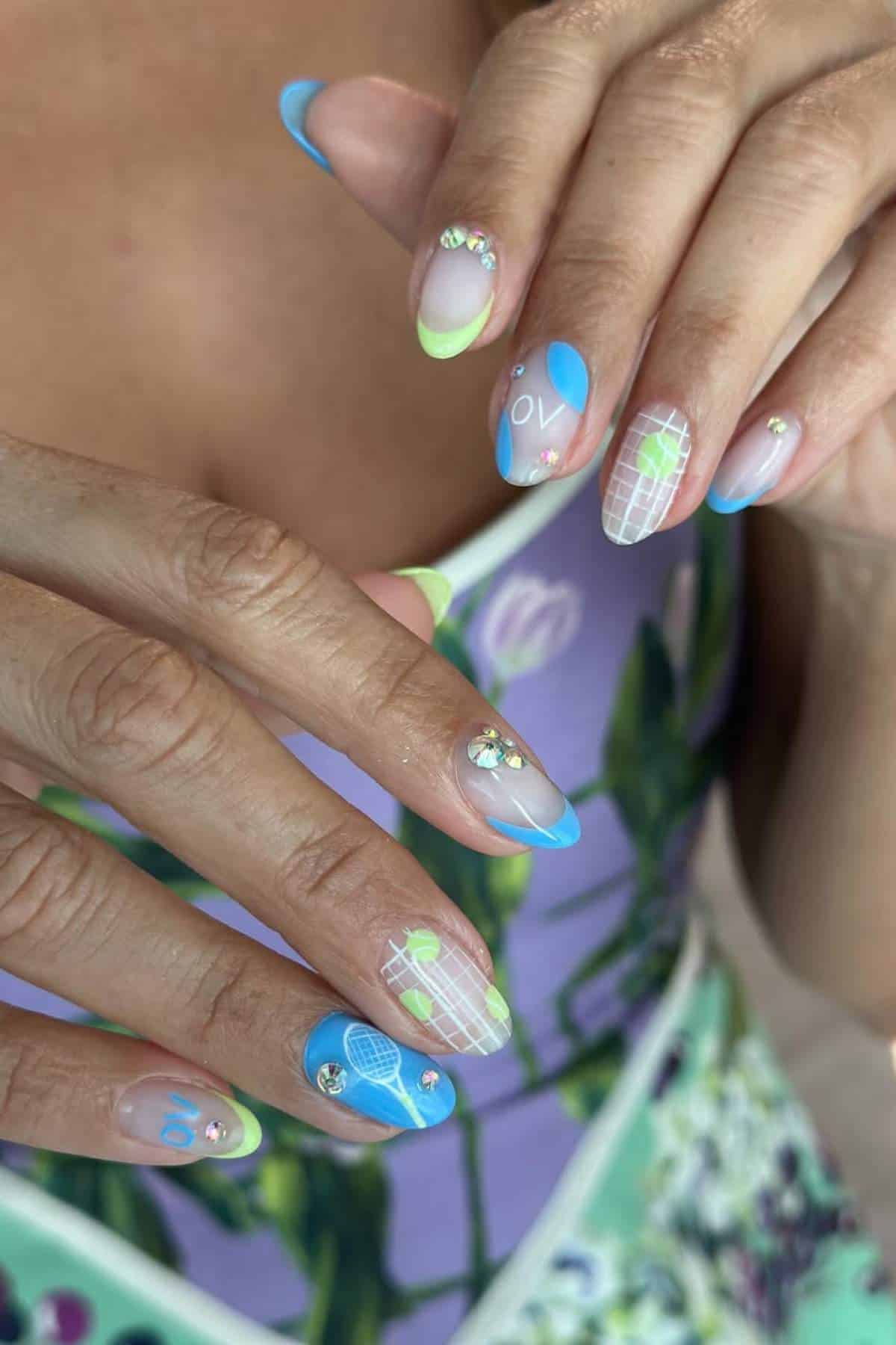 A close-up of hands with nail art featuring blue, green, and white tennis-themed designs, rhinestone accents, and a floral dress in the background.