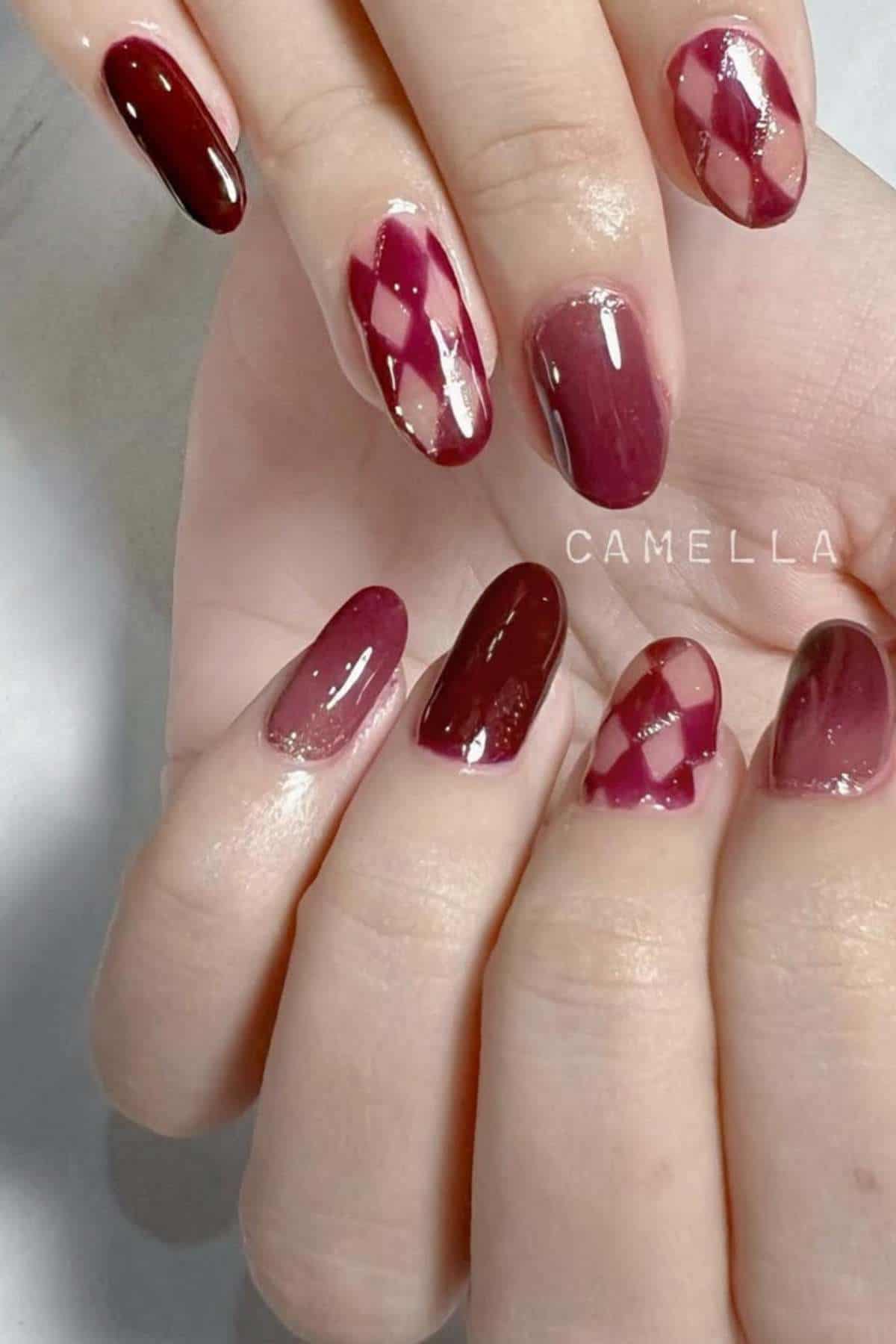 Hands with almond-shaped nails painted in varying shades of burgundy, featuring solid colors and diamond-shaped argyle patterns on some nails.