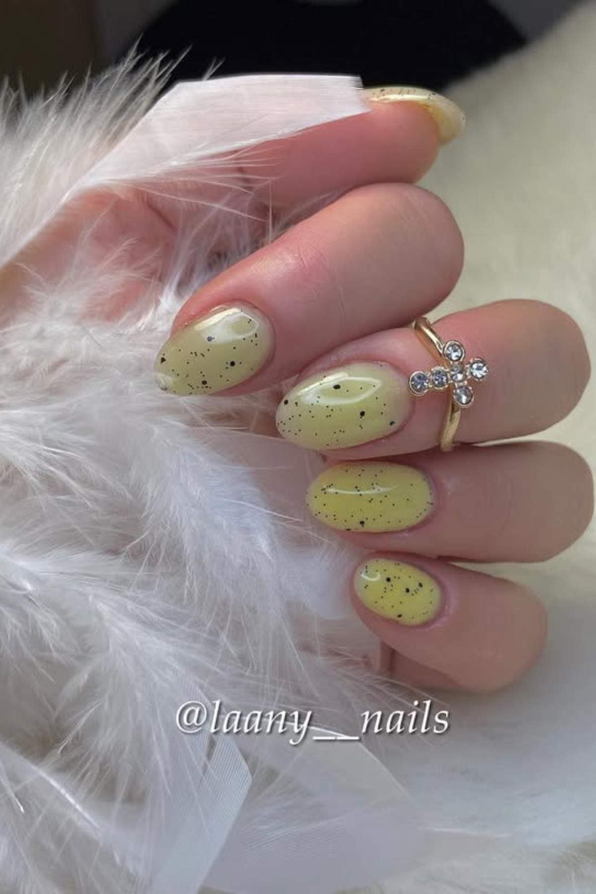 A hand with short, oval-shaped nails painted light yellow with black speckles, holding a white feather; wearer has a gold ring with rhinestones.