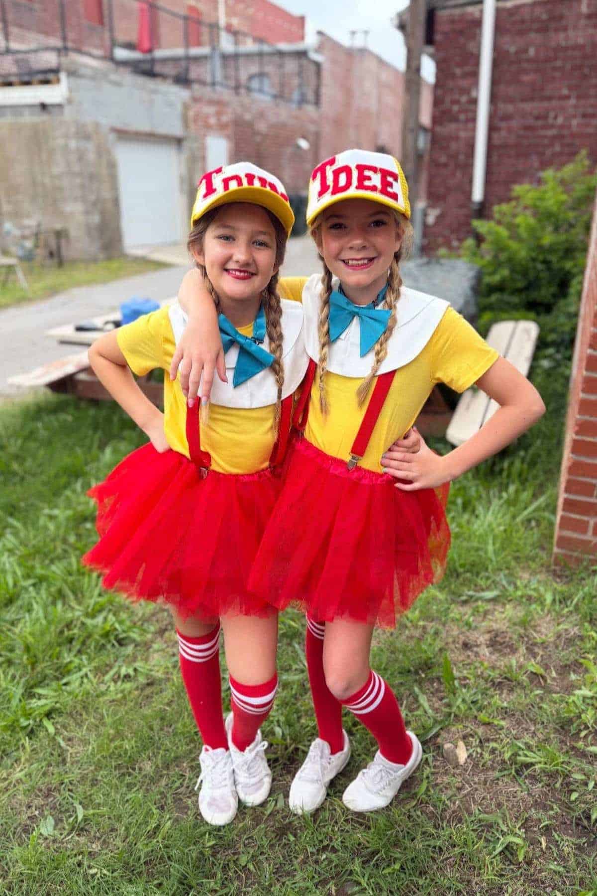 Two young girls stand outdoors wearing matching yellow shirts, red tutus, white collars, suspenders, knee-high socks, and caps labeled "DEE" and "DUM.