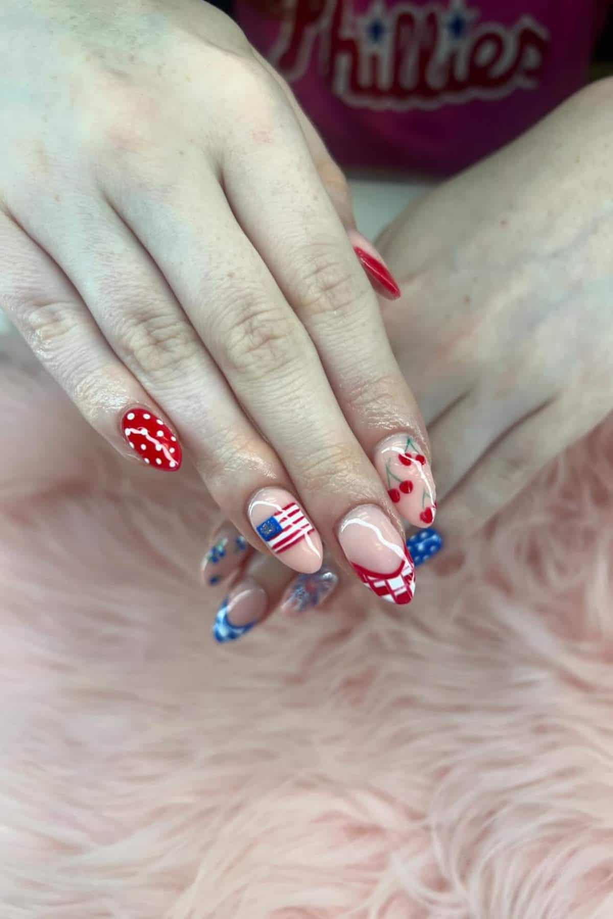Hands with manicured nails featuring red, white, and blue designs, including stars, stripes, cherries, and checkered patterns, resting on a pink furry surface.