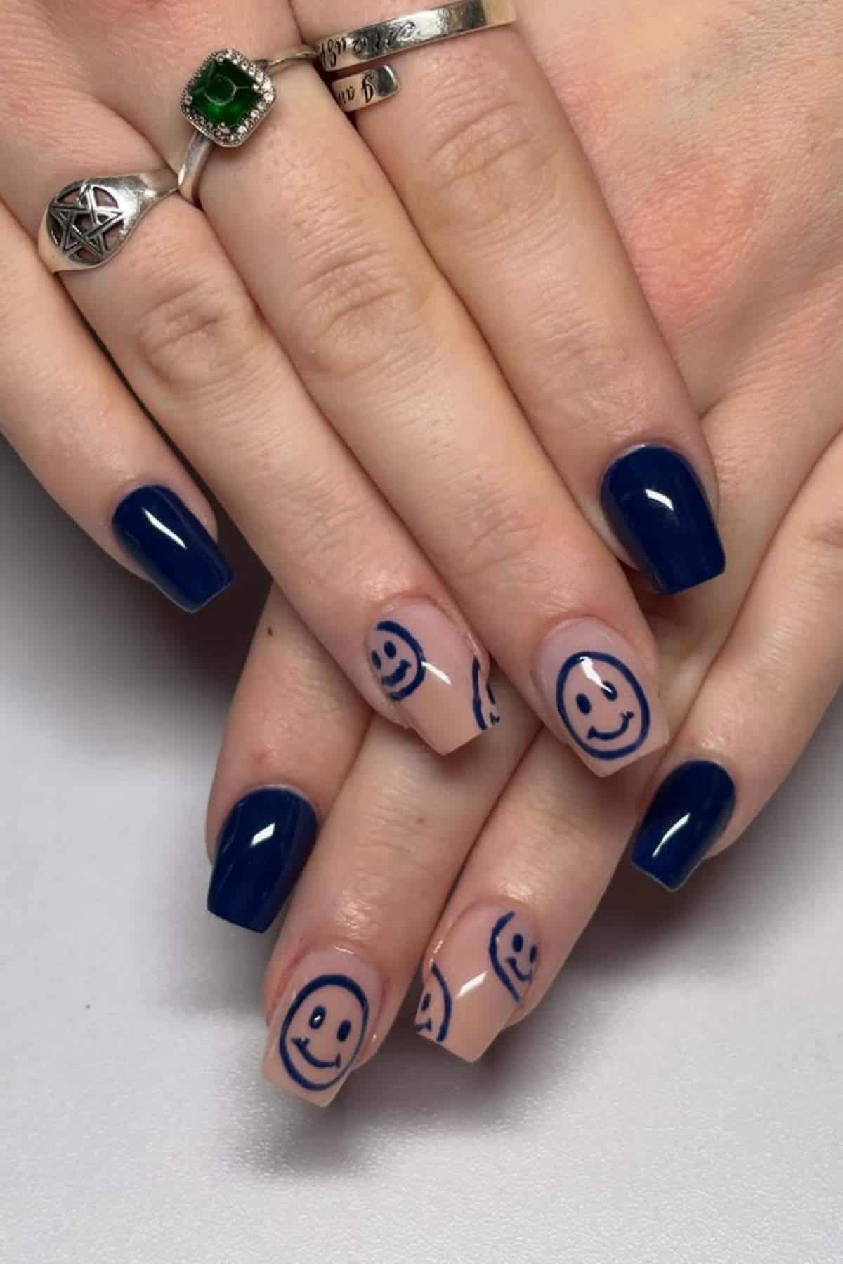 Hands with manicured nails; some nails are painted dark blue, while others are nude with blue smiley face designs. The person is wearing silver rings.