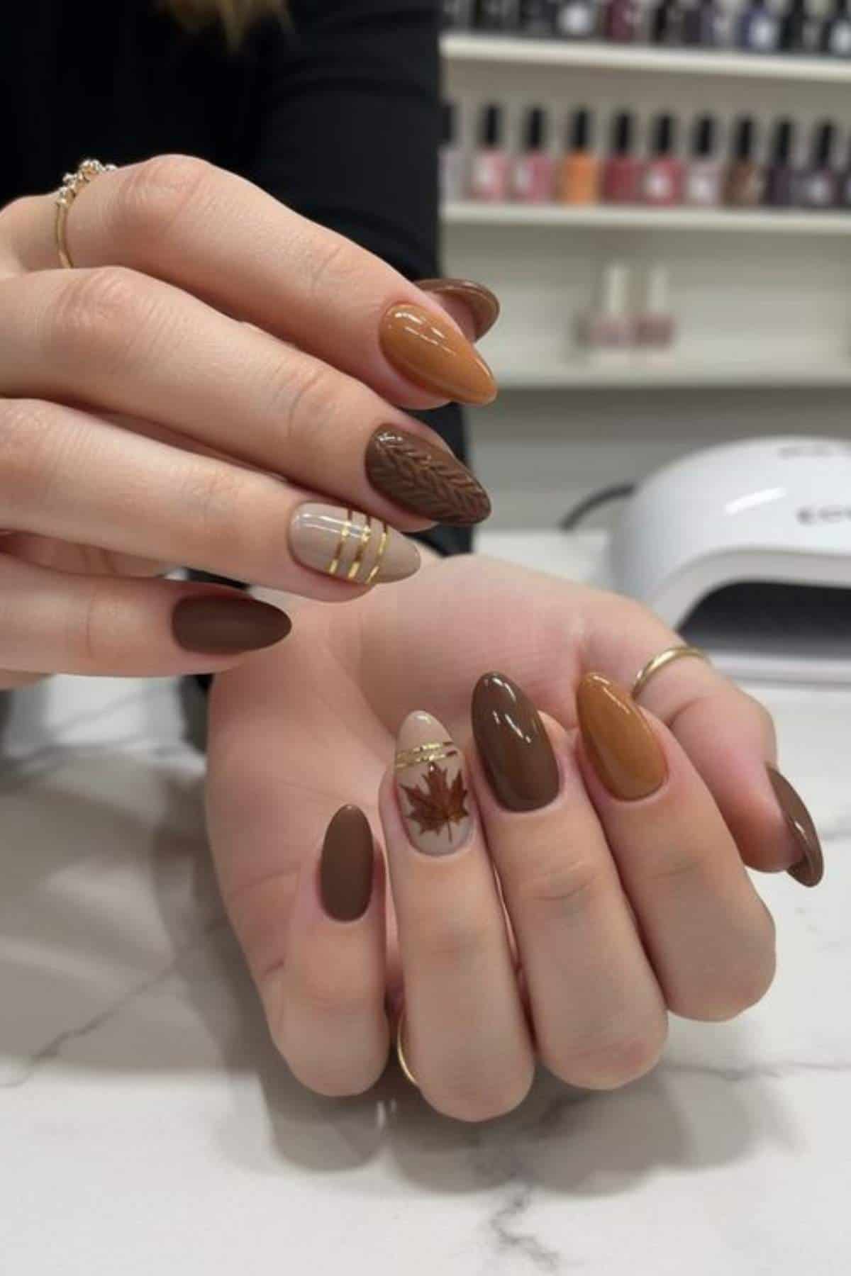A hand with almond-shaped nails painted in various shades of brown, featuring gold stripes, a leaf pattern, and a maple leaf design, shown on a marble surface.