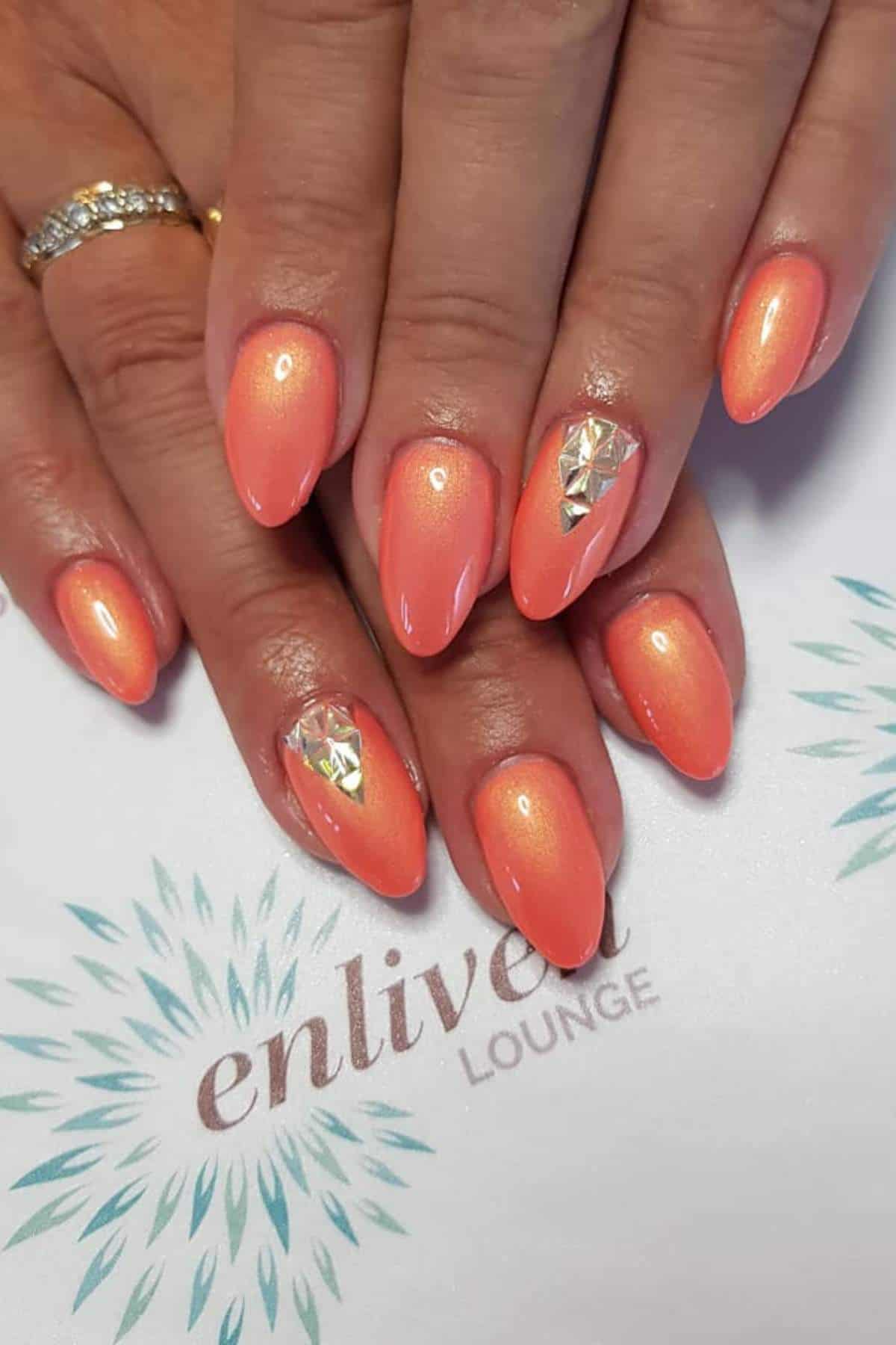 Hands with almond-shaped, coral-orange nails featuring a glossy finish and geometric silver accents on the ring fingernails, resting on an "enliven LOUNGE" card.