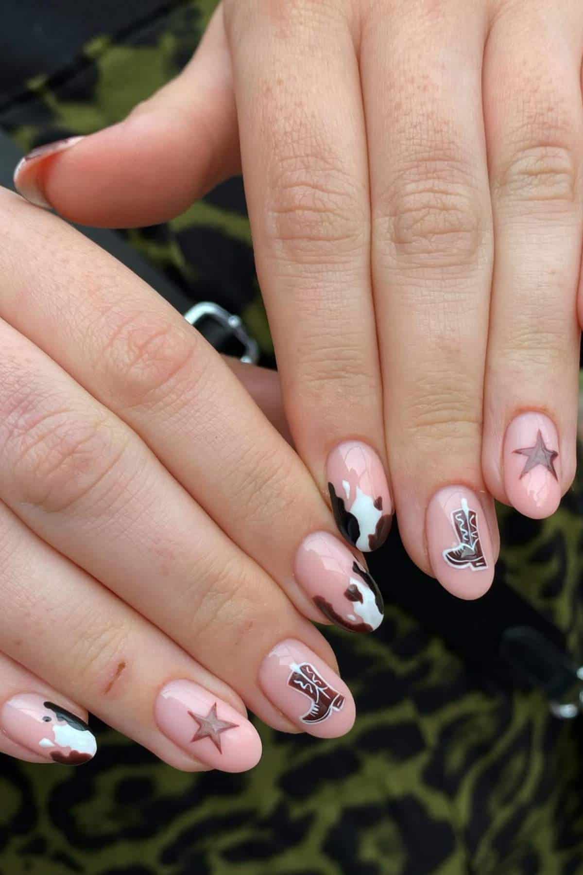 Close-up of hands with a nude-pink manicure featuring black and white cow print, silver sheriff star, and cowboy boot designs on fingernails.