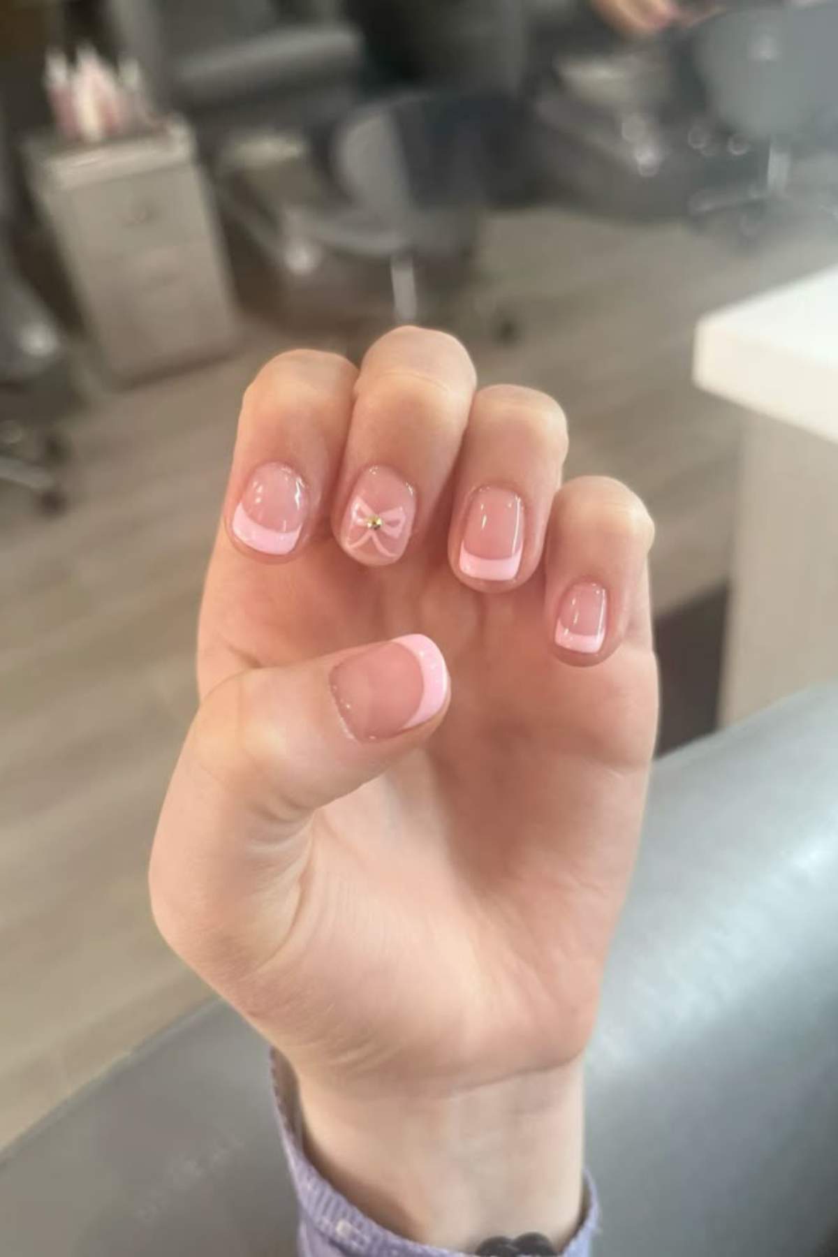 A hand with a light pink French manicure; one nail features a small gold bow decoration.