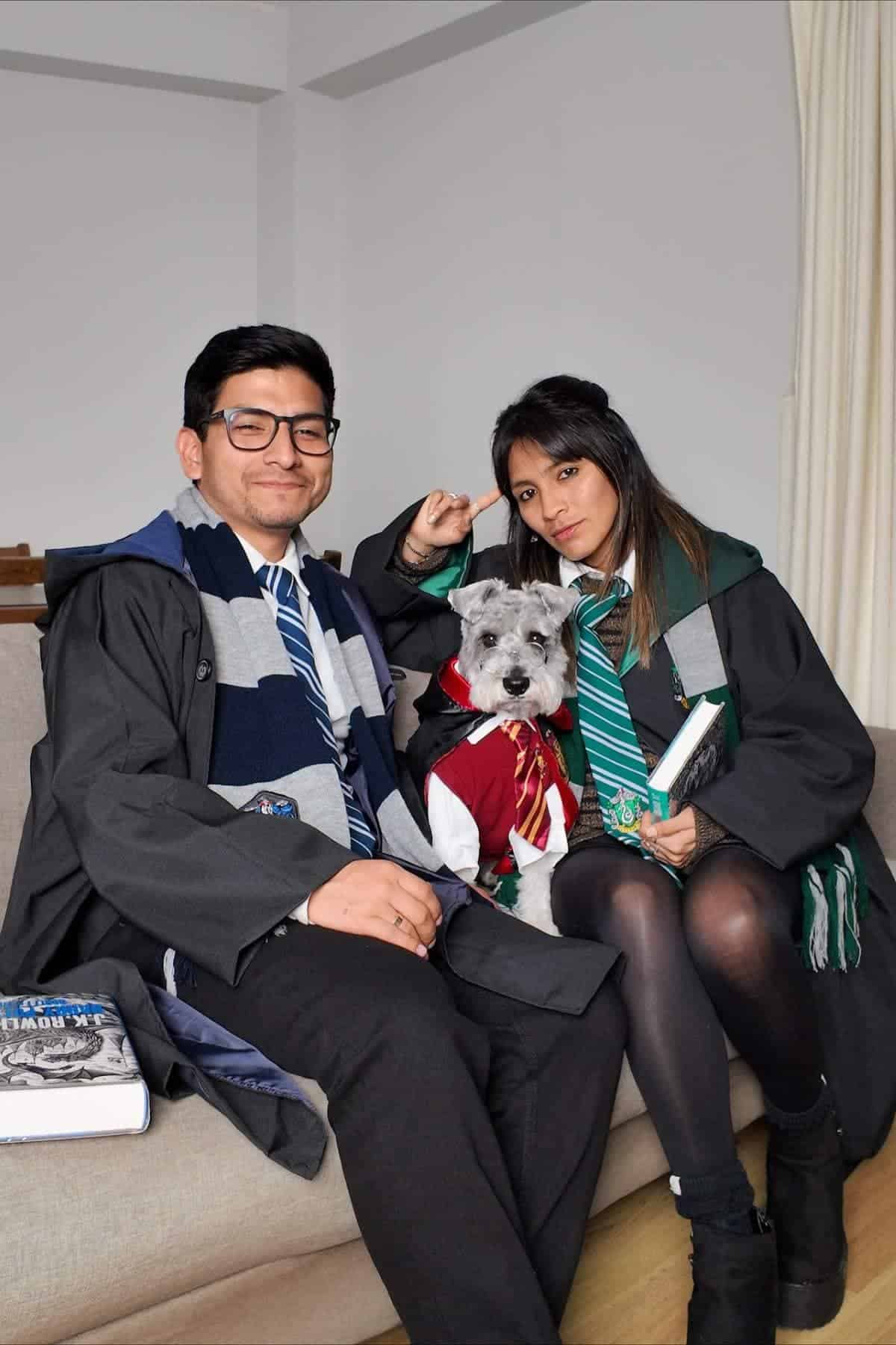 Two people dressed in Harry Potter-themed costumes sit on a couch with a small dog in a Gryffindor outfit. One person holds a book and the other holds a wand.