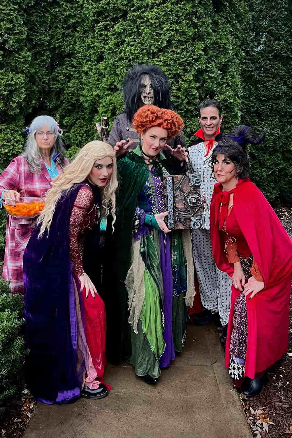 Six adults in colorful costumes pose outdoors, dressed as characters from the movie "Hocus Pocus," with greenery in the background.