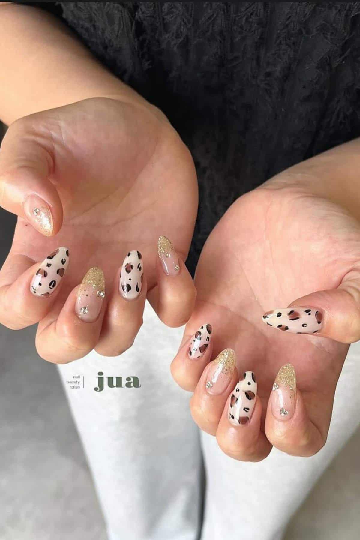 Hands with almond-shaped nails featuring a mix of gold glitter tips and white nails with black and brown leopard print design.