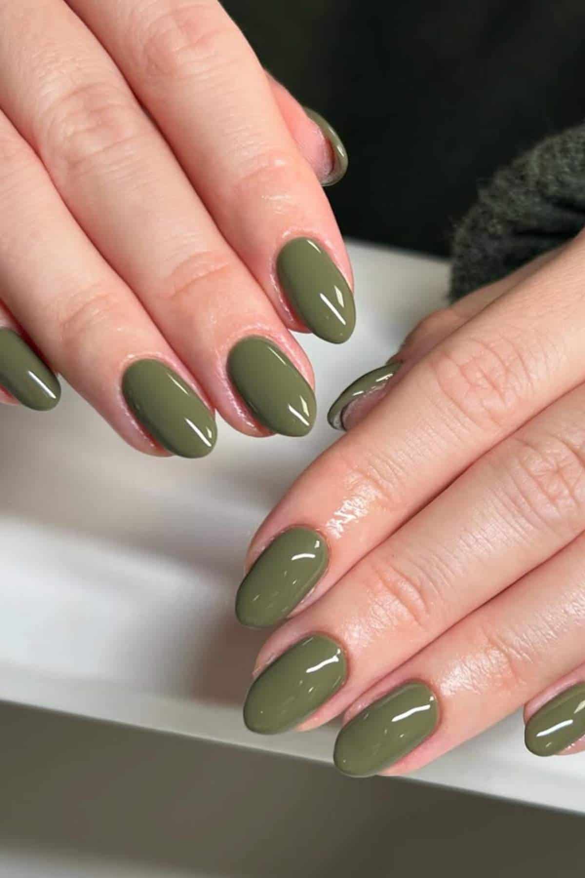 Close-up of hands with oval-shaped nails painted in glossy olive green nail polish.