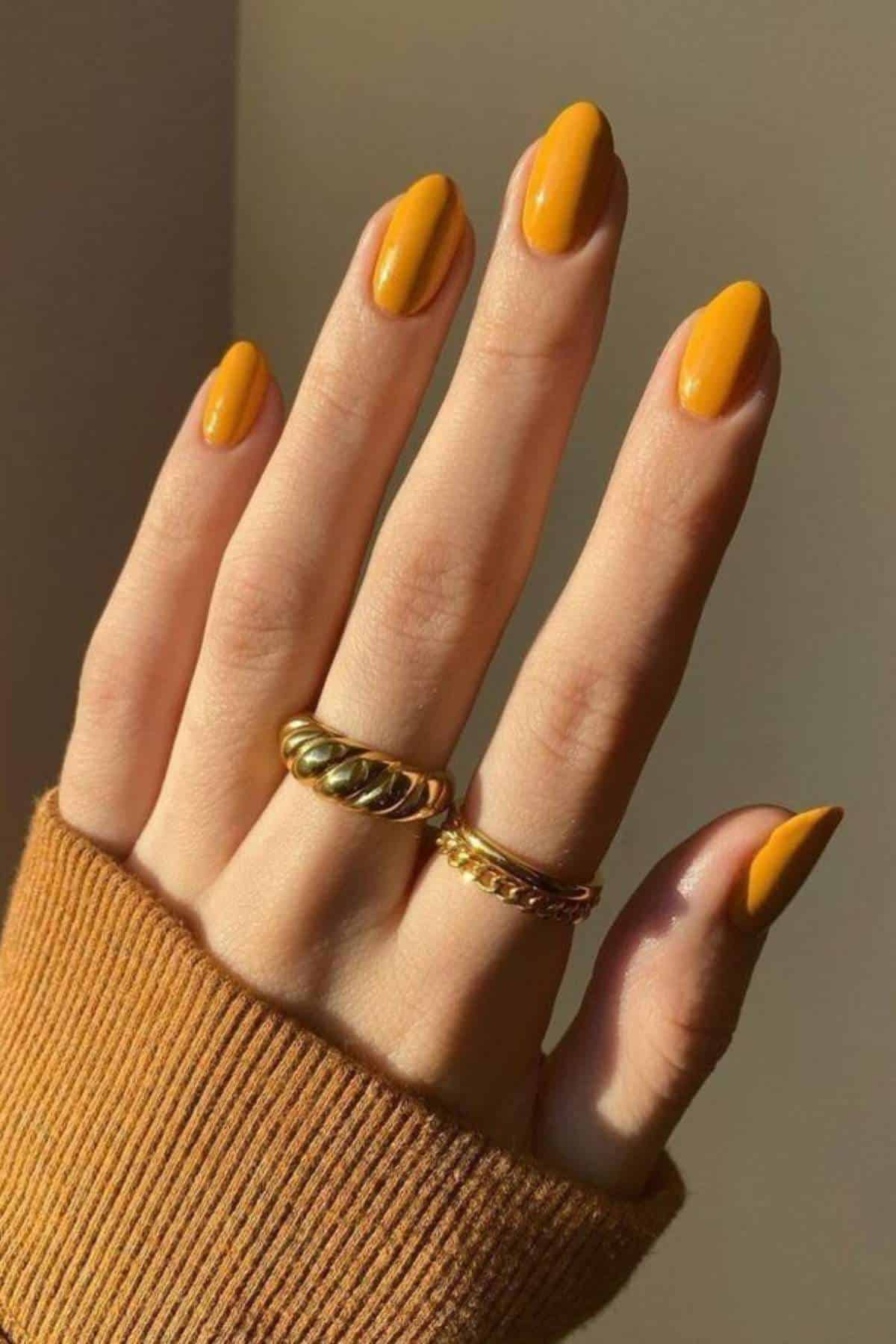 A hand with yellow-orange painted nails, wearing two gold rings, is shown against a neutral background. The person is wearing a ribbed brown sleeve.
