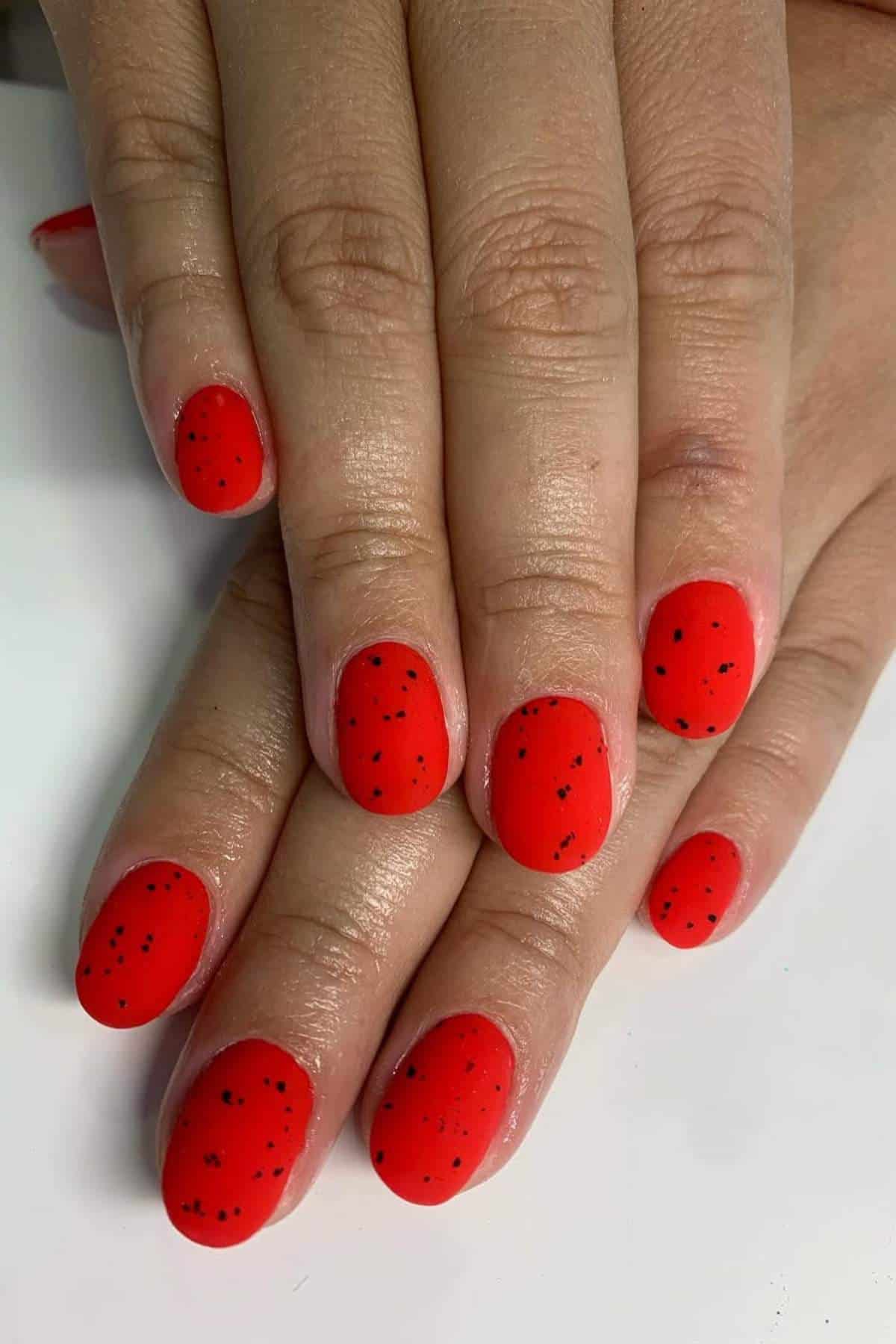 Hands with short, oval-shaped nails painted bright red with small black speckles, resembling a watermelon effect, resting on a white surface.