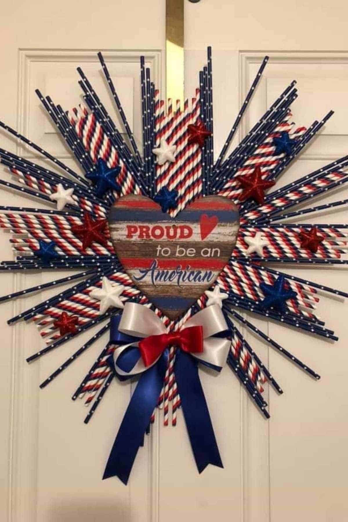 A patriotic wreath with red, white, and blue straws, stars, a heart-shaped sign reading "Proud to be an American," and a red, white, and blue ribbon bow.
