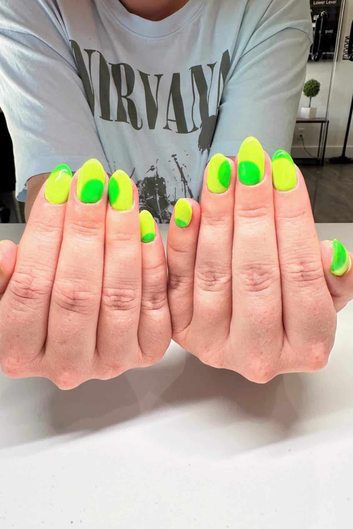 Person with light skin showing both hands; fingernails painted neon yellow-green with darker green dots. Person is wearing a faded Nirvana T-shirt. Indoor setting.