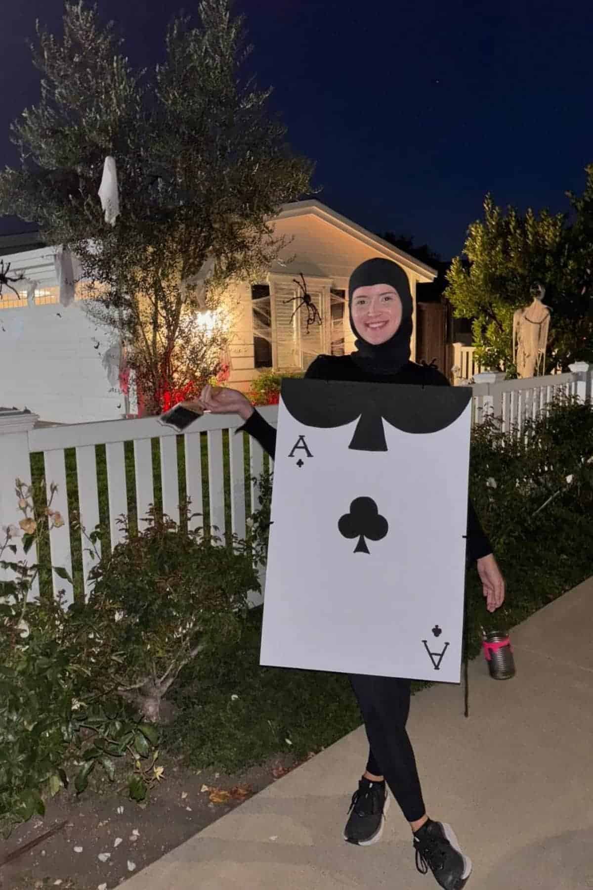 Person wearing an Ace of Clubs playing card costume stands on a sidewalk in front of a white house at night, holding a drink container and smiling.