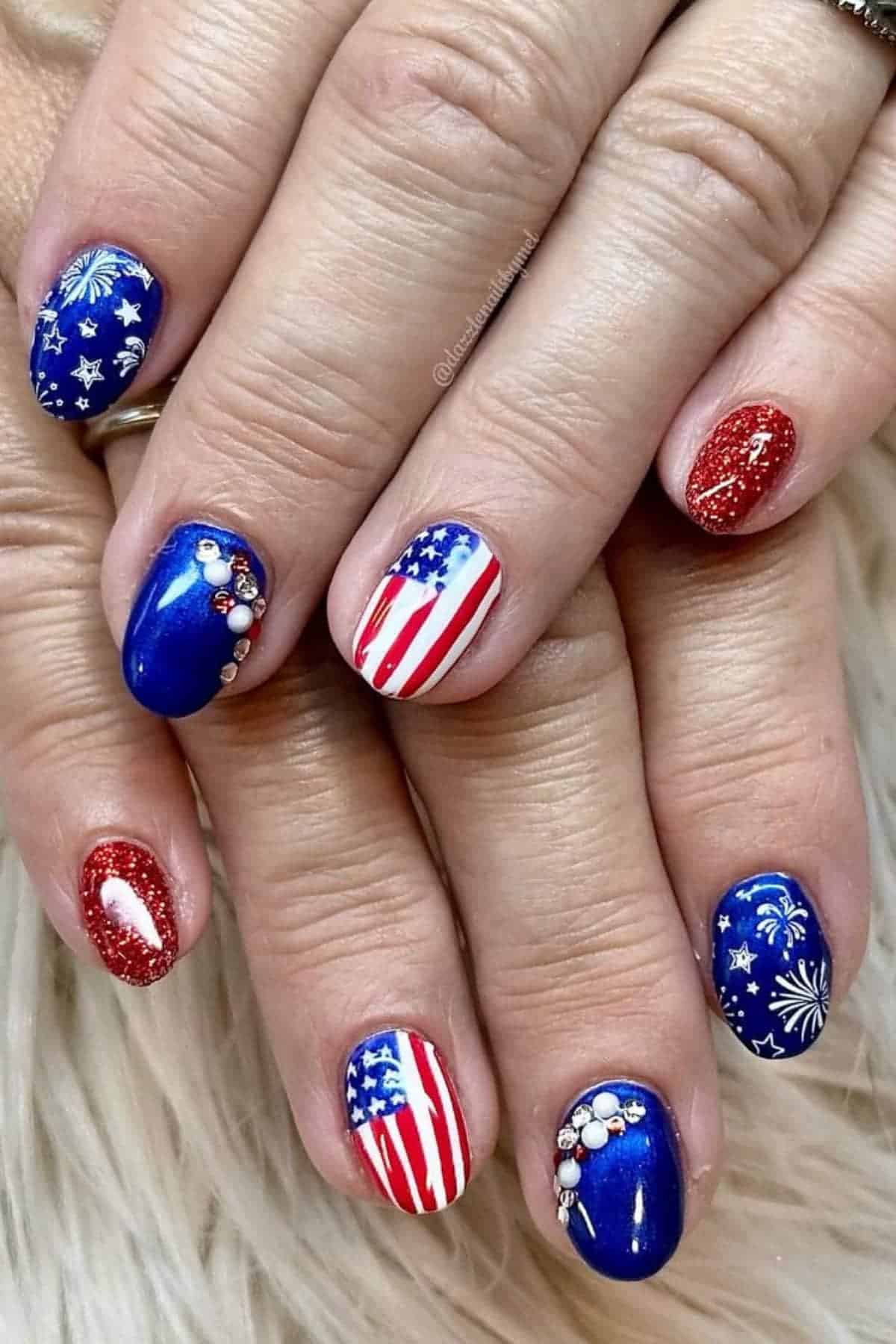 Hands with manicured nails featuring American flag designs, blue with white fireworks, red glitter, and rhinestone accents.
