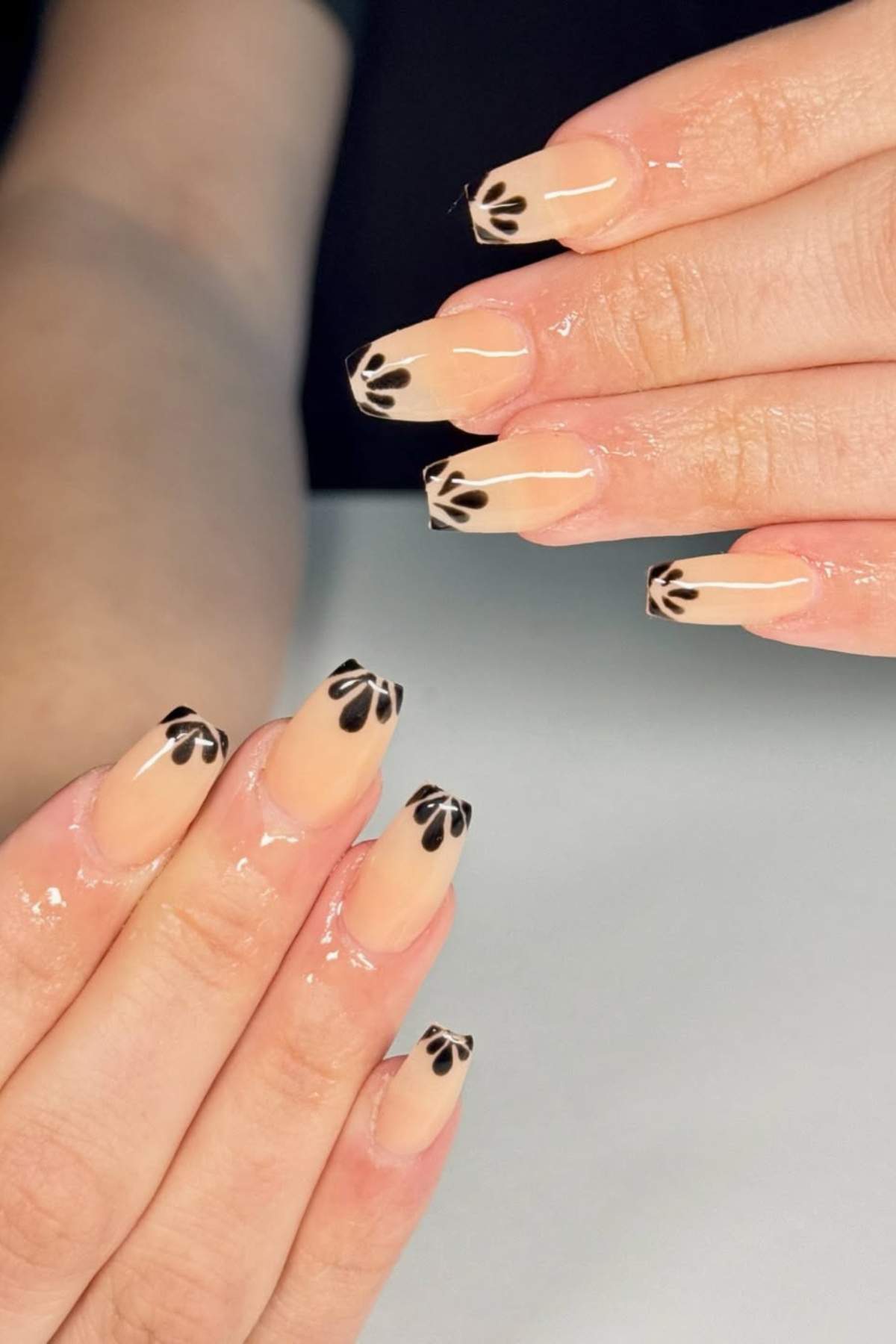Hands with long, square nude nails featuring black petal-like designs on the tips, displayed under a soft light.