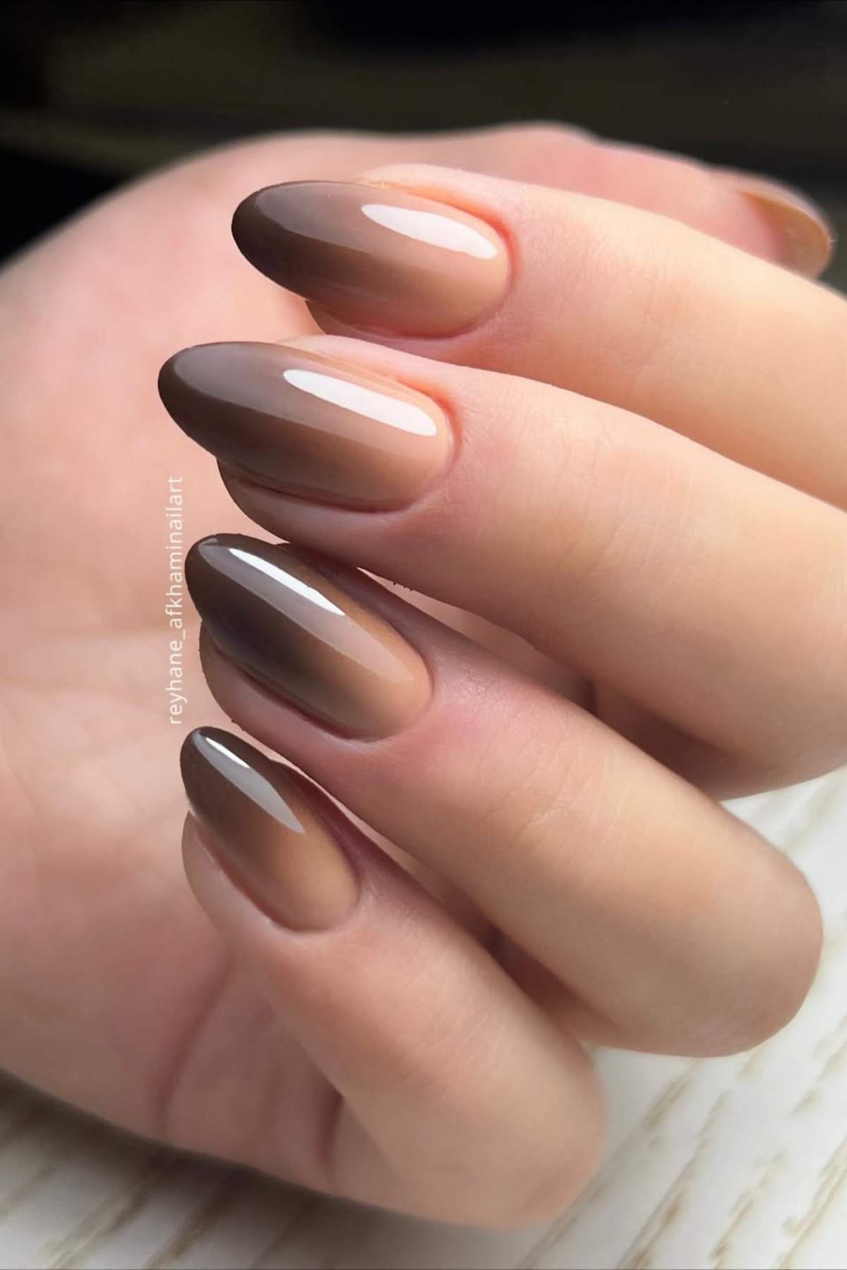 A hand with almond-shaped nails featuring a glossy brown gradient ombre manicure.