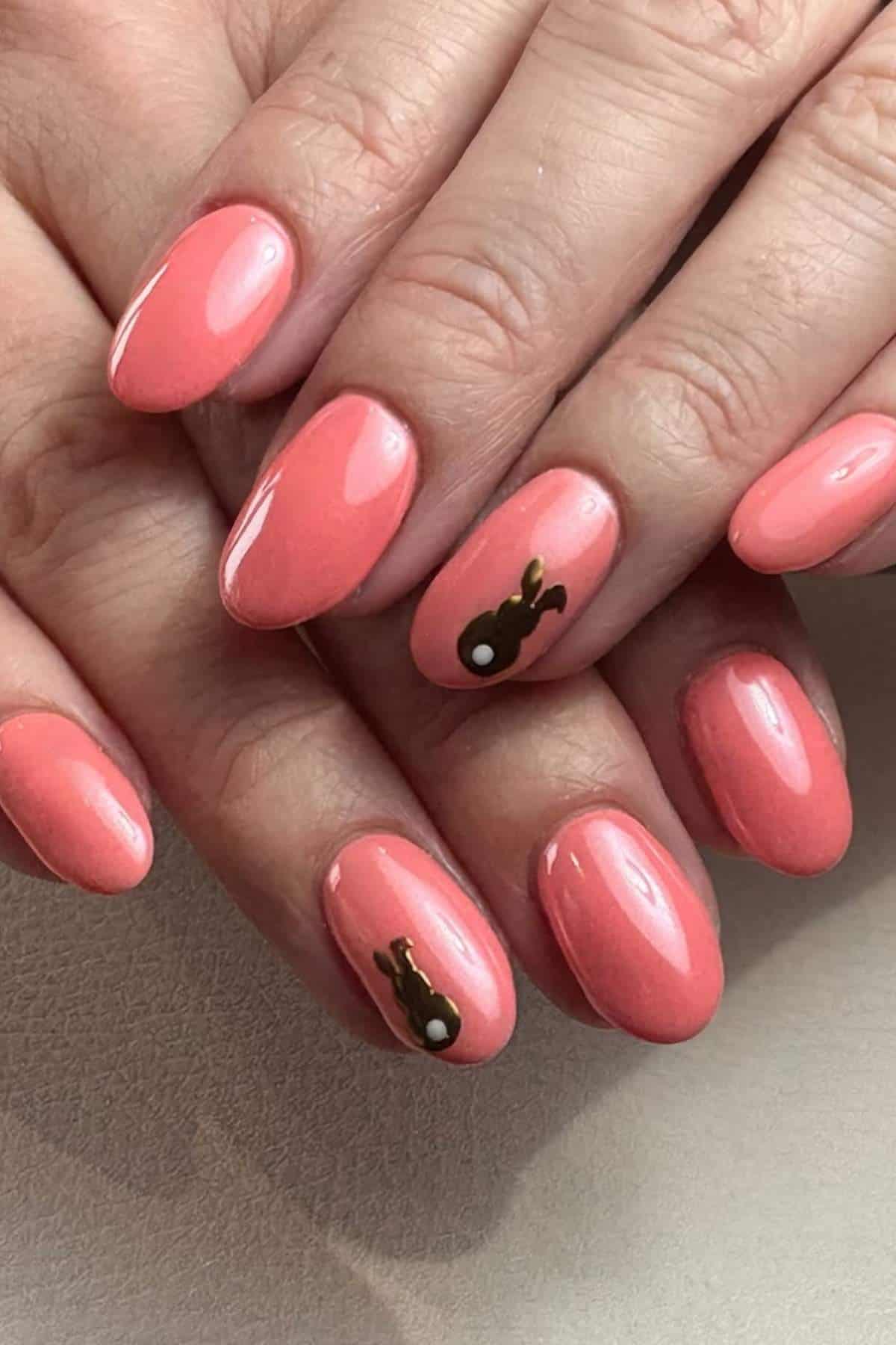 Hands with oval-shaped, glossy coral pink nails; ring fingers have a small brown bunny design with a white tail as nail art accent.
