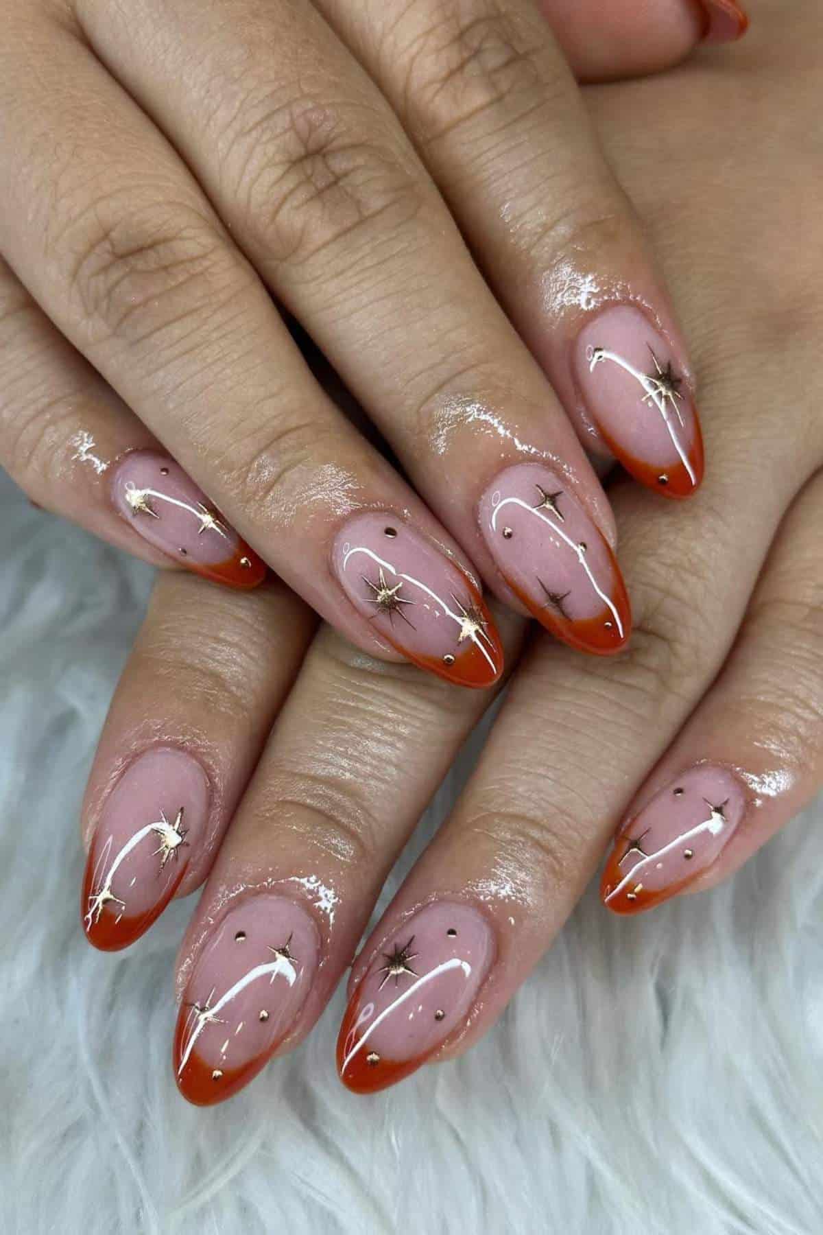 Hands with almond-shaped nails featuring orange tips, gold star decals, white line designs, and small black dots on a light pink base, resting on a white textured surface.