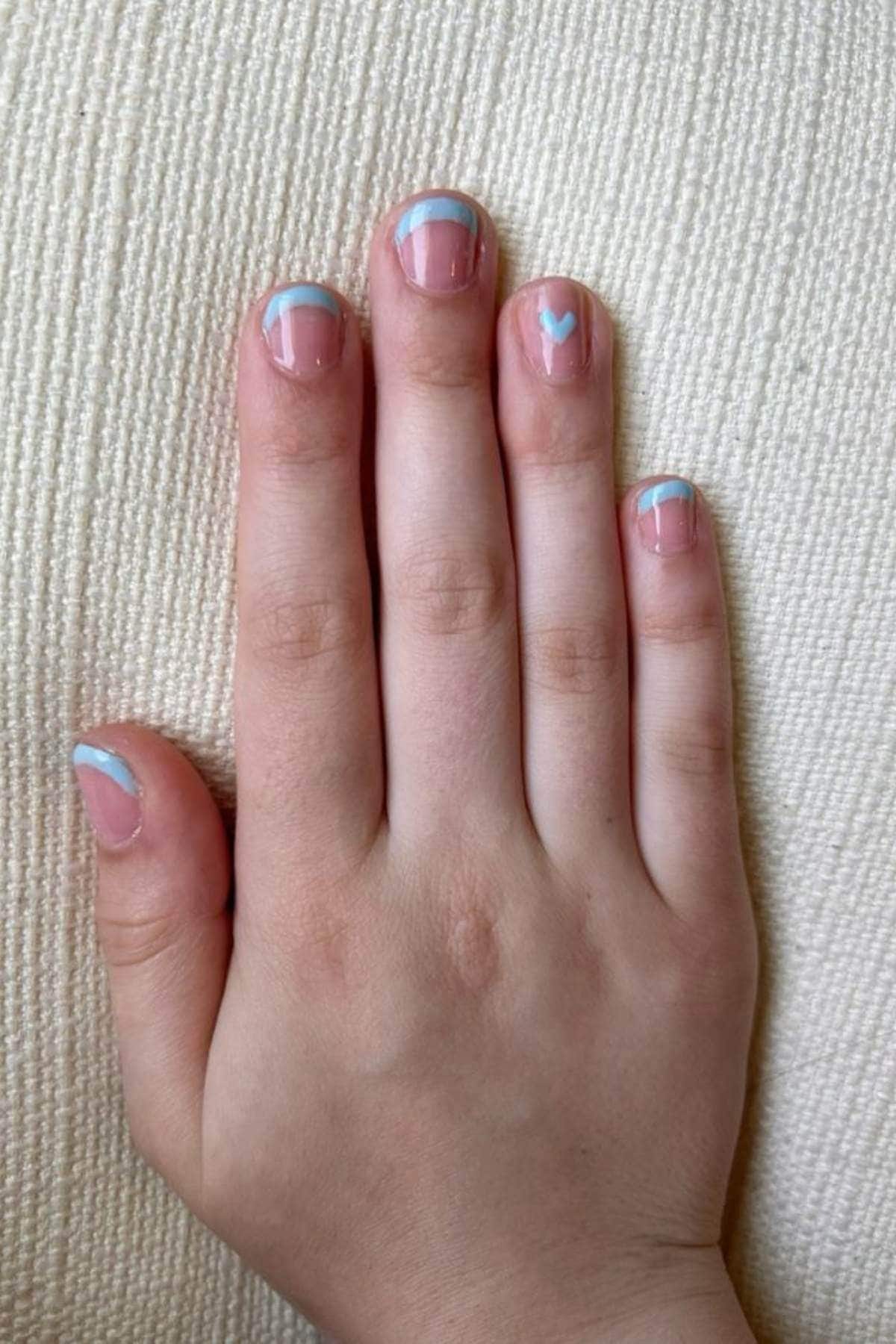 A hand with short nails painted with light blue and white tips, and a small white heart on the middle fingernail, resting on a textured cream fabric.