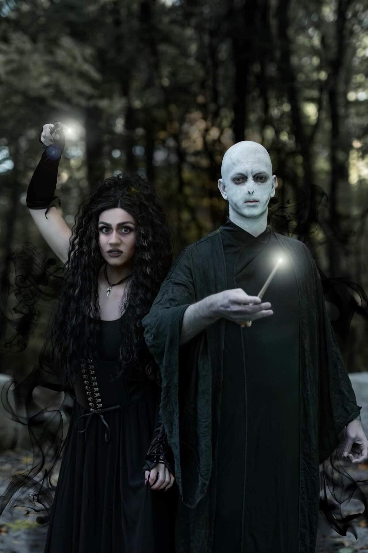 Two people in dark costumes, one with long black hair and the other bald with pale skin, stand in a forest, each holding a wand emitting light.