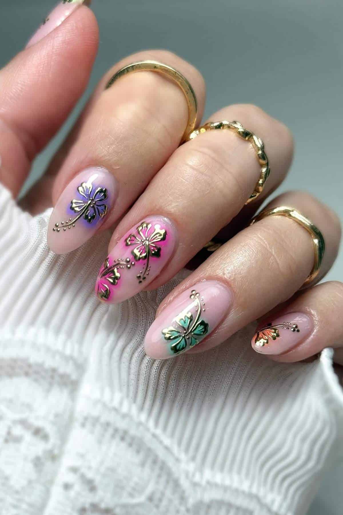 A hand with almond-shaped nails painted in pastel colors, decorated with metallic butterfly nail art and small rhinestones, wearing gold rings.