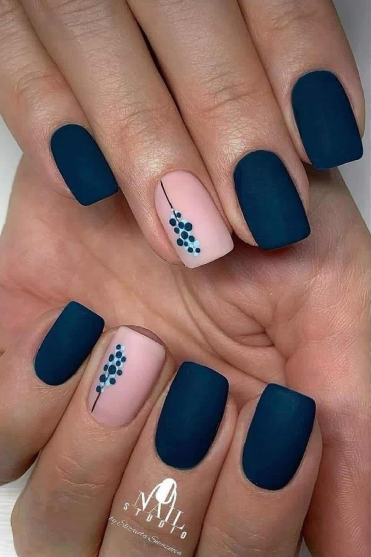 Hands with short, square-shaped nails painted matte navy blue, except two accent nails in nude with a simple blue dot floral design.