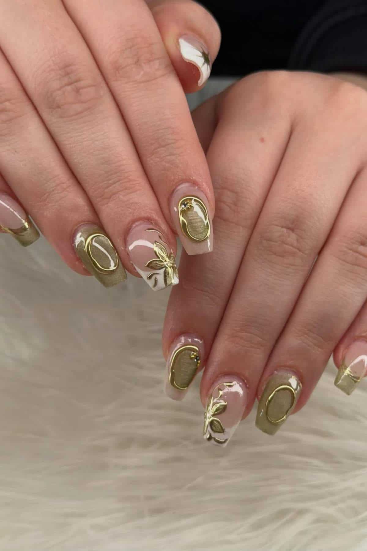 Two hands with manicured nails featuring olive green polish, gold 3D accents, and white floral designs on a light fur surface.