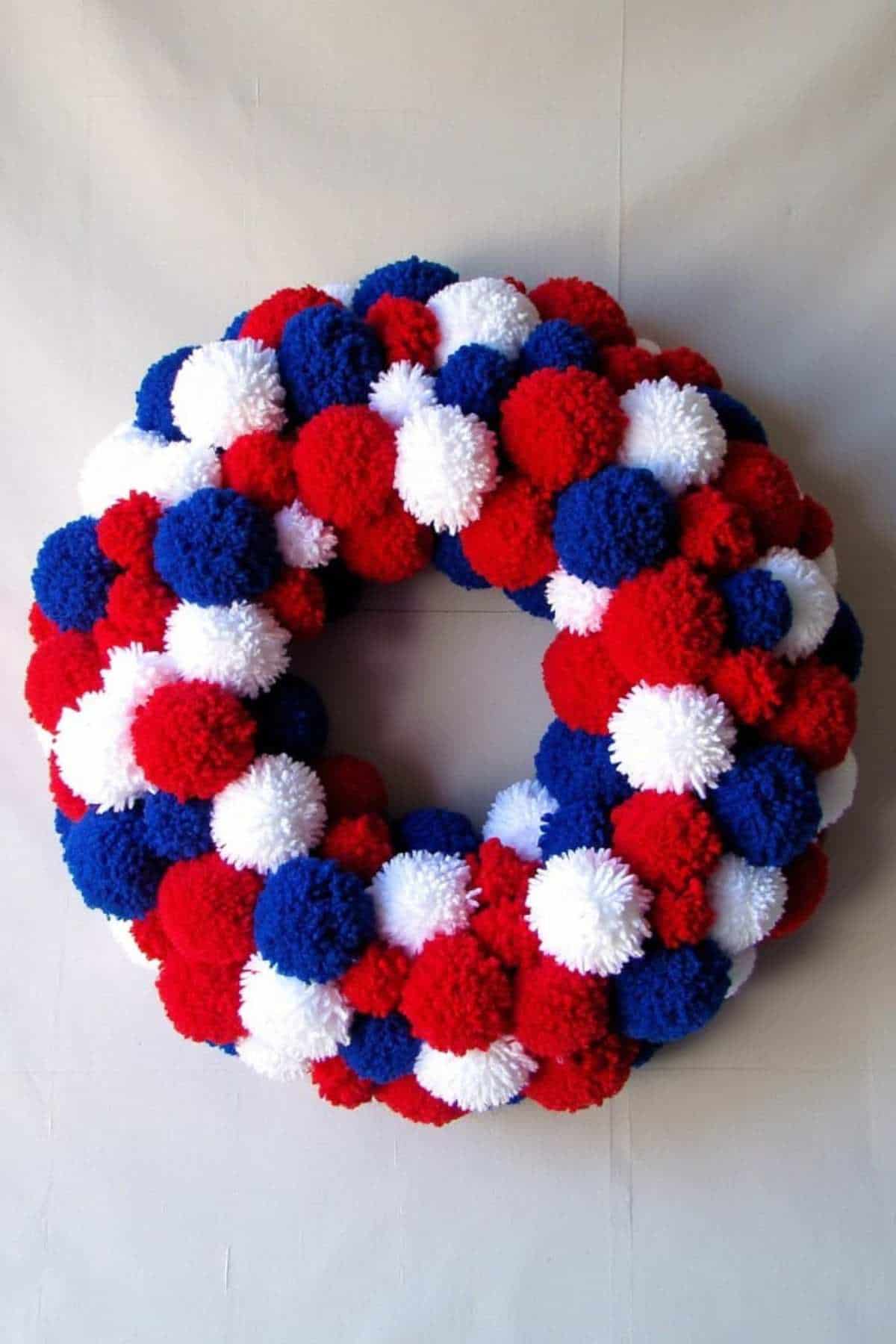 A wreath made of red, white, and blue pom-poms arranged in a circular pattern against a plain background.